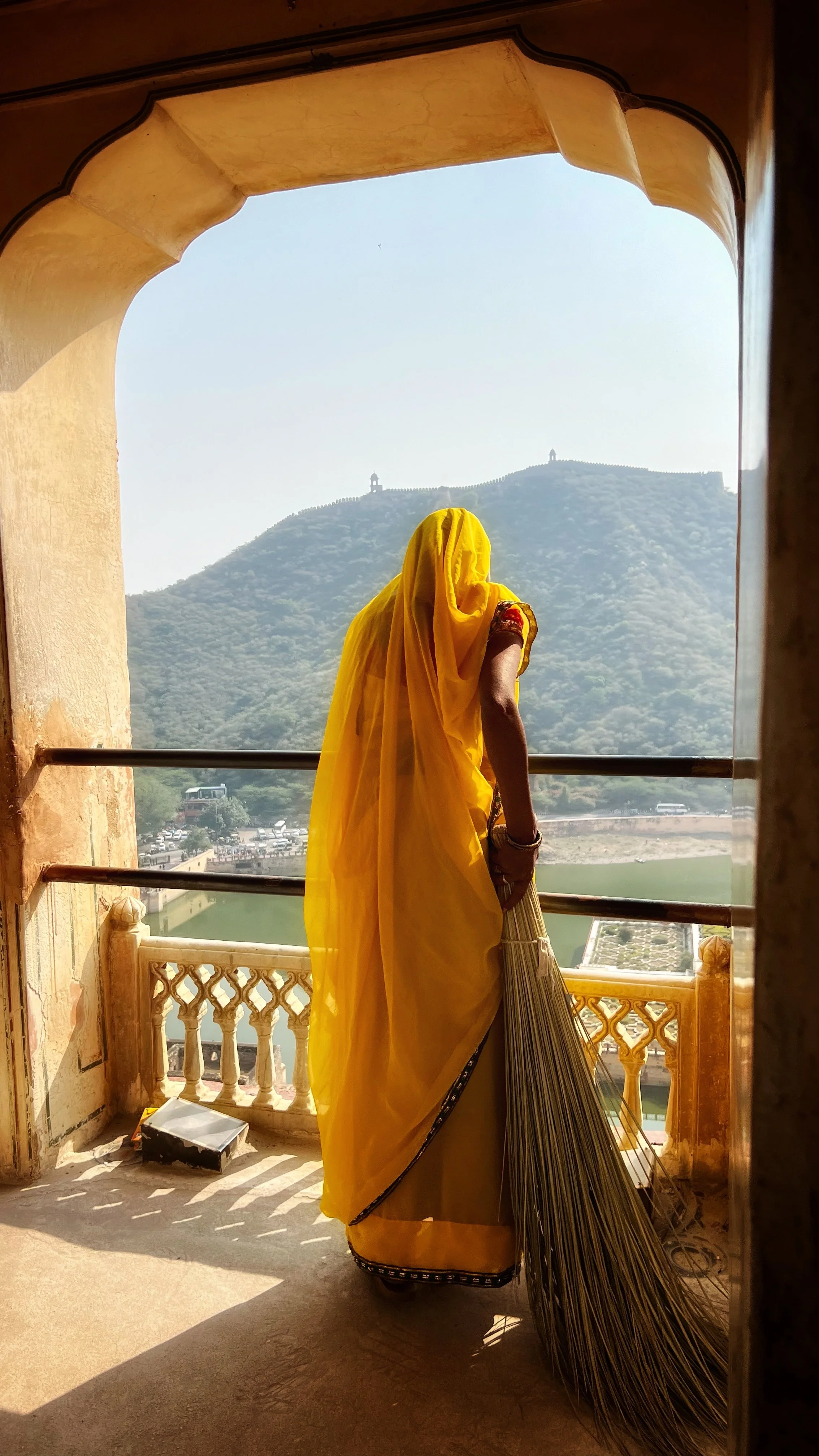 A woman wearing a yellow sari stands on a balcony, looking at a hill with small structures on top.