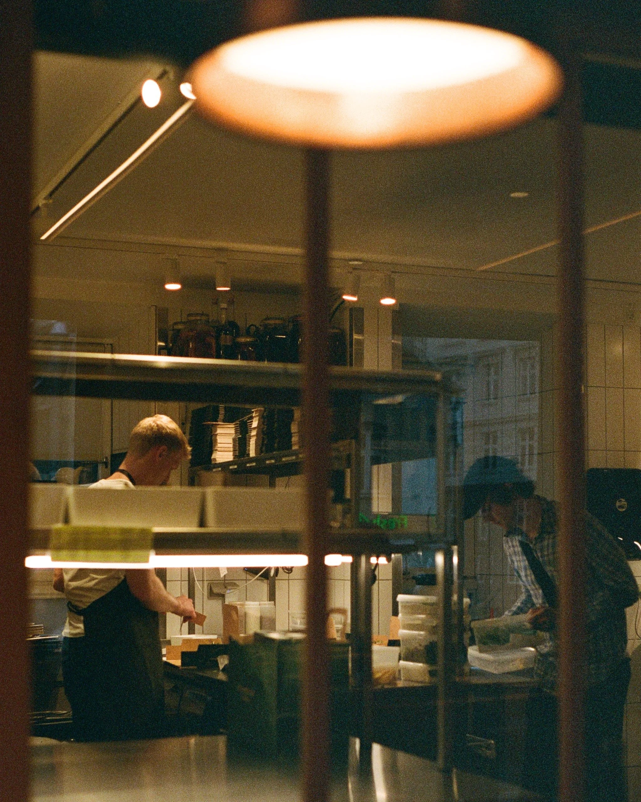 A man working behind a kitchen counter, preparing food, in a restaurant setting with shelves of kitchen equipment and shelves of books or similar stacked in the background, seen through a window with a grid of vertical and horizontal bars.