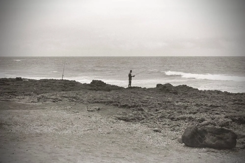 A person fishing with a fishing rod on rocky shoreline near the ocean, in black and white.