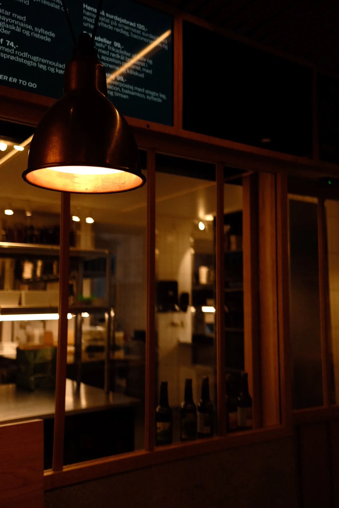 Indoor restaurant or bar interior with warm lighting, featuring a hanging lamp, wooden dividing panels, and a digital menu screen in the background.