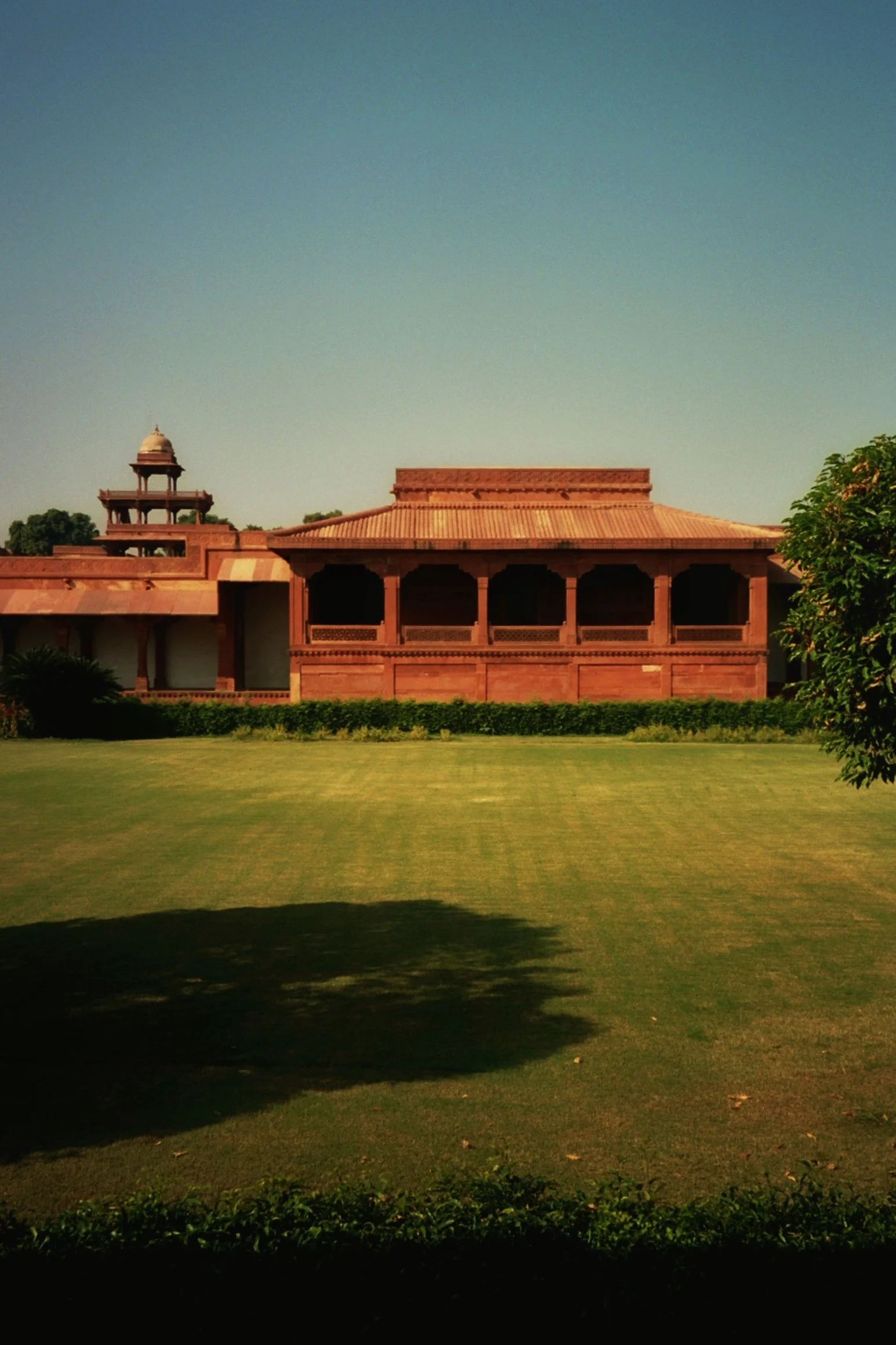 A large historic building made of red sandstone, with a central open veranda and a small structure with domes on the left side, set in a lush green garden under a clear blue sky.