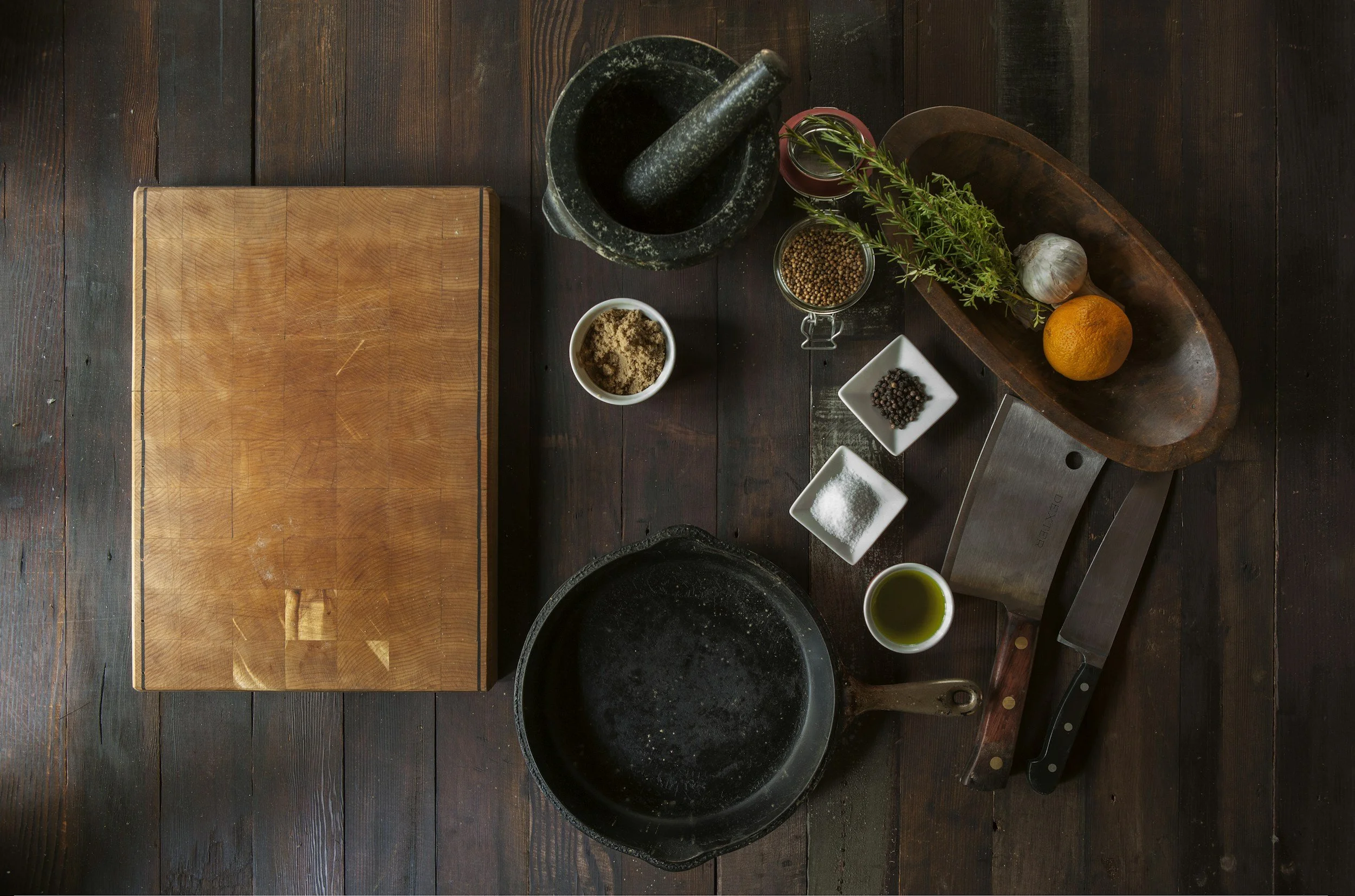Top-down view of a vintage wooden kitchen setup with a cutting board, kitchen tools, herbs, garlic, lemon, and spices on a dark wooden table.