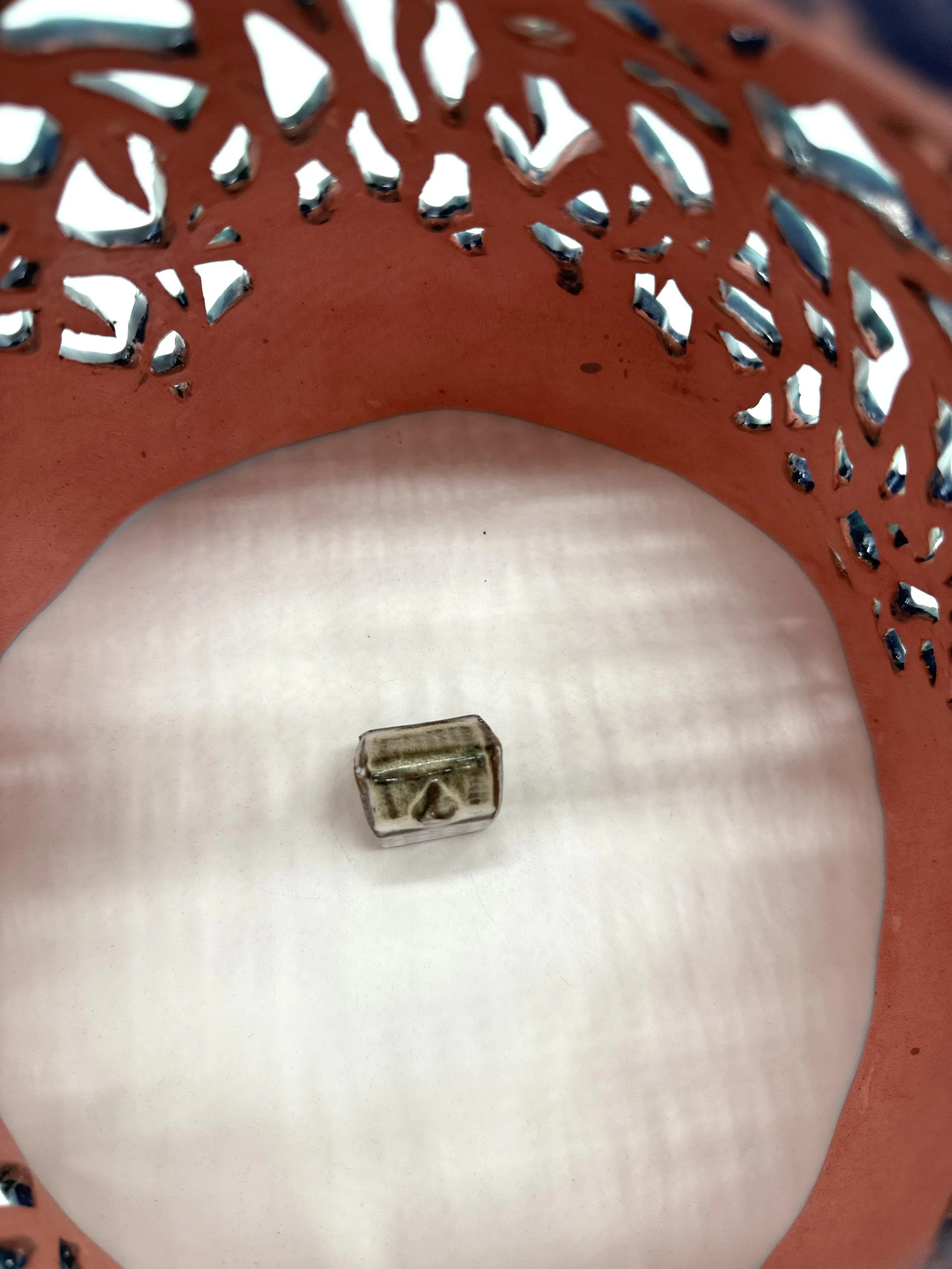 A small glass cube with a metallic key inside, placed on a white surface underneath an orange, decorative, perforated metal object.