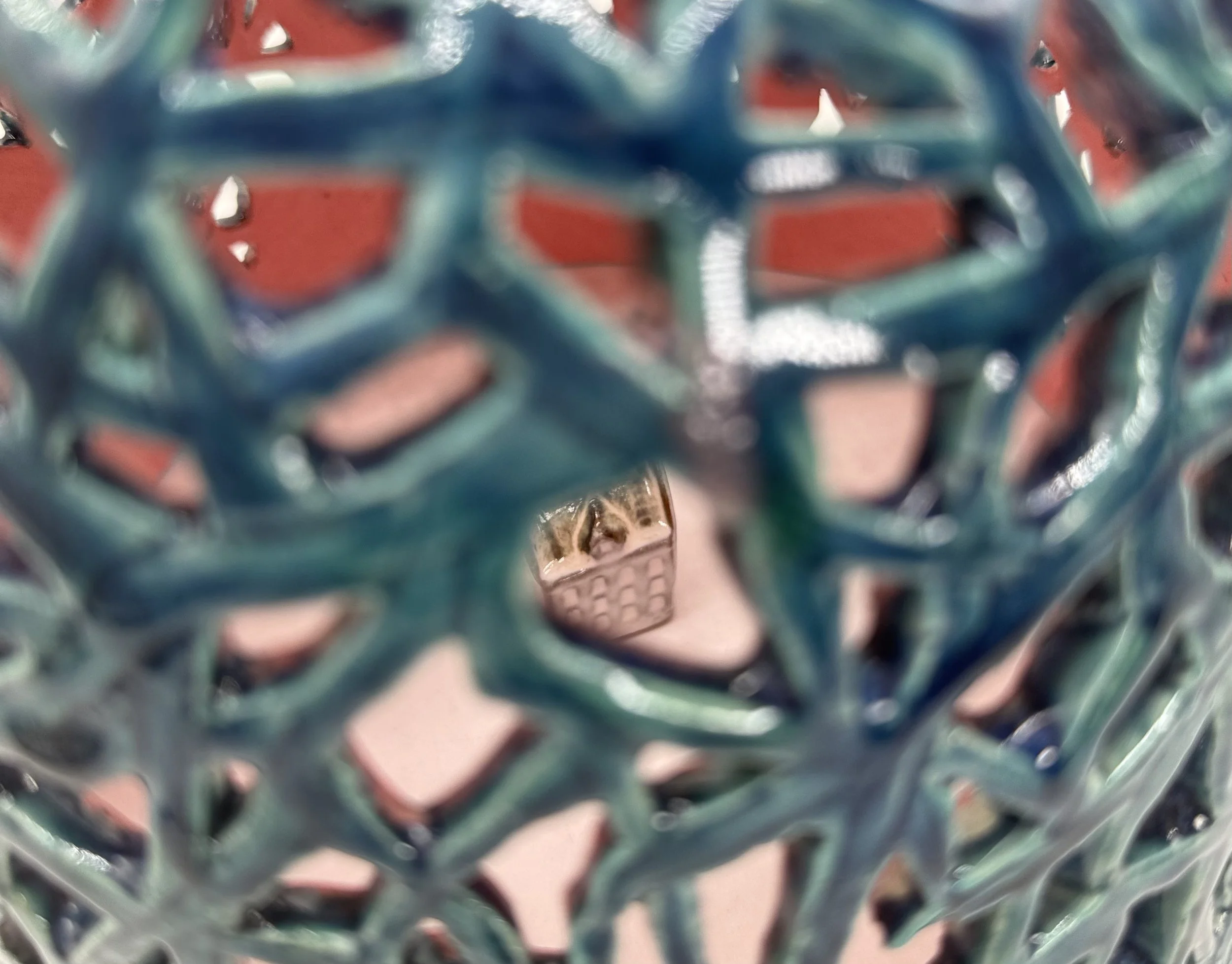 Close-up view of a decorative metal sphere with a blue-green patina, featuring an intricate open geometric pattern, with a blurred red background.