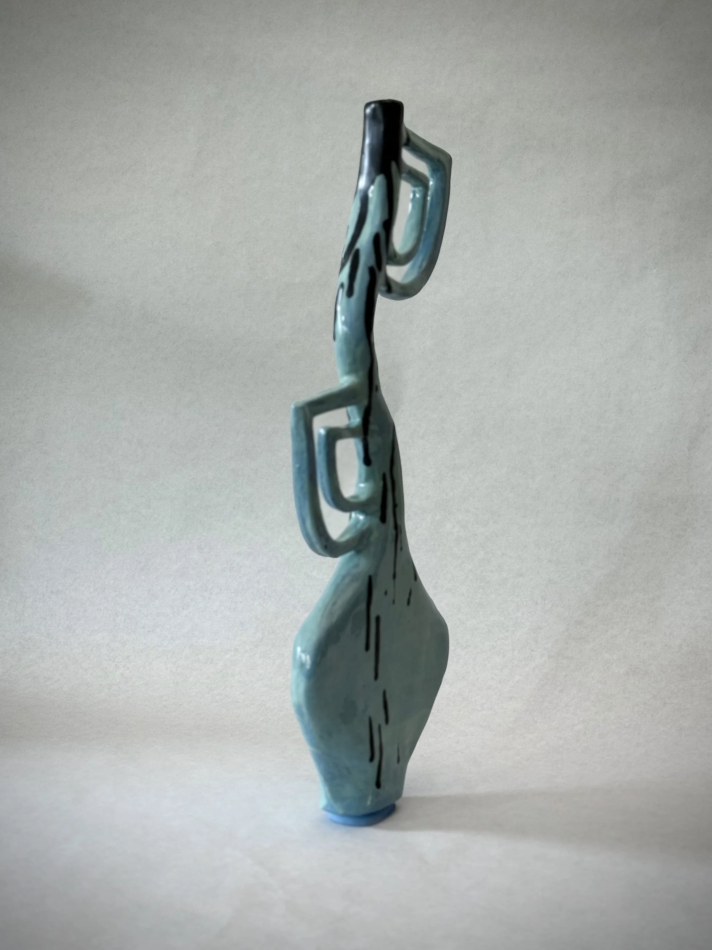 Ceramic sculpture of a stylized, elongated face with abstract features, painted in shades of blue and black, mounted on a white surface.