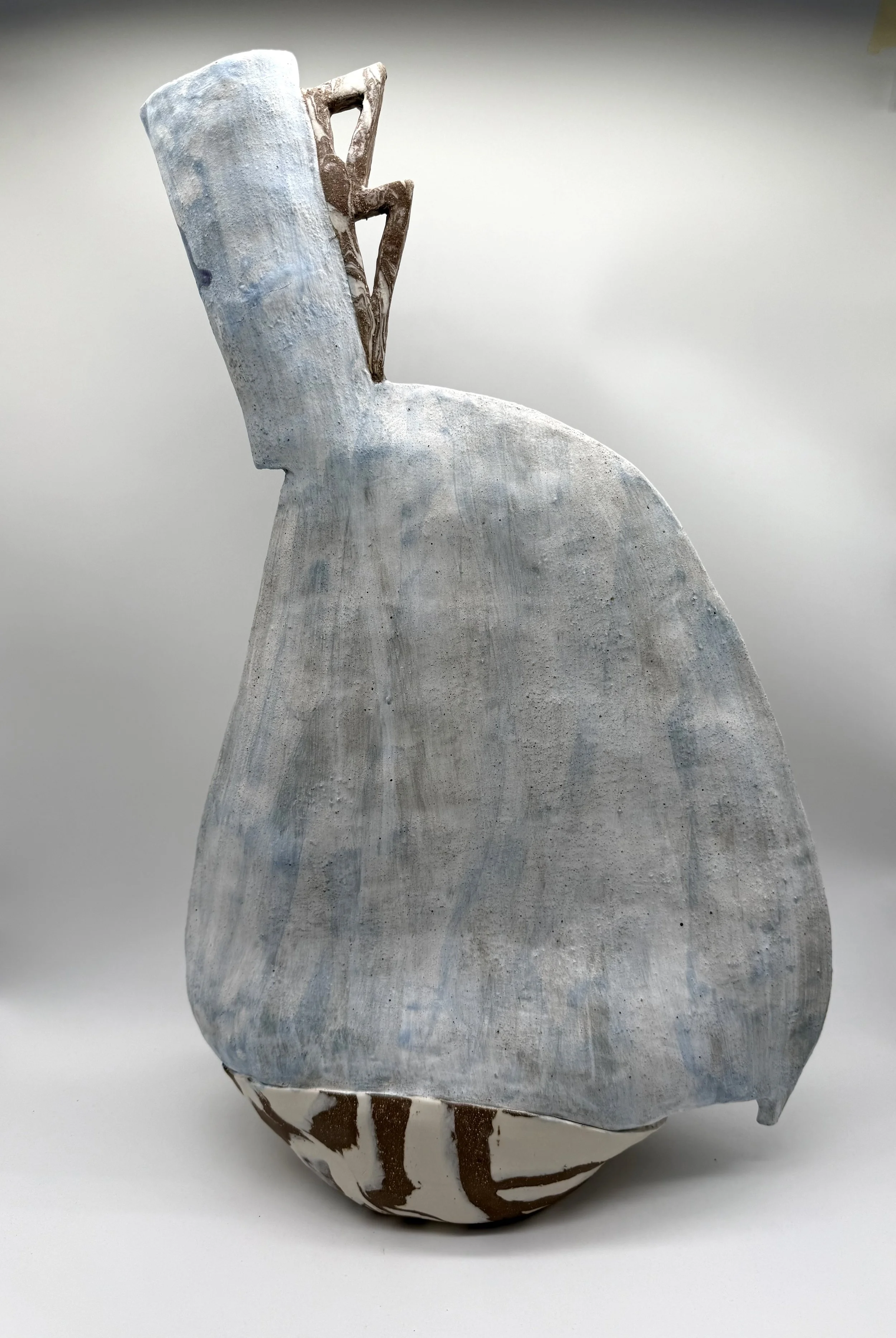 A ceramic sculpture resembling a stylized animal, with a large, smooth, light blue body and a smaller head with abstract features. The sculpture has brown and white striped elements at its base.