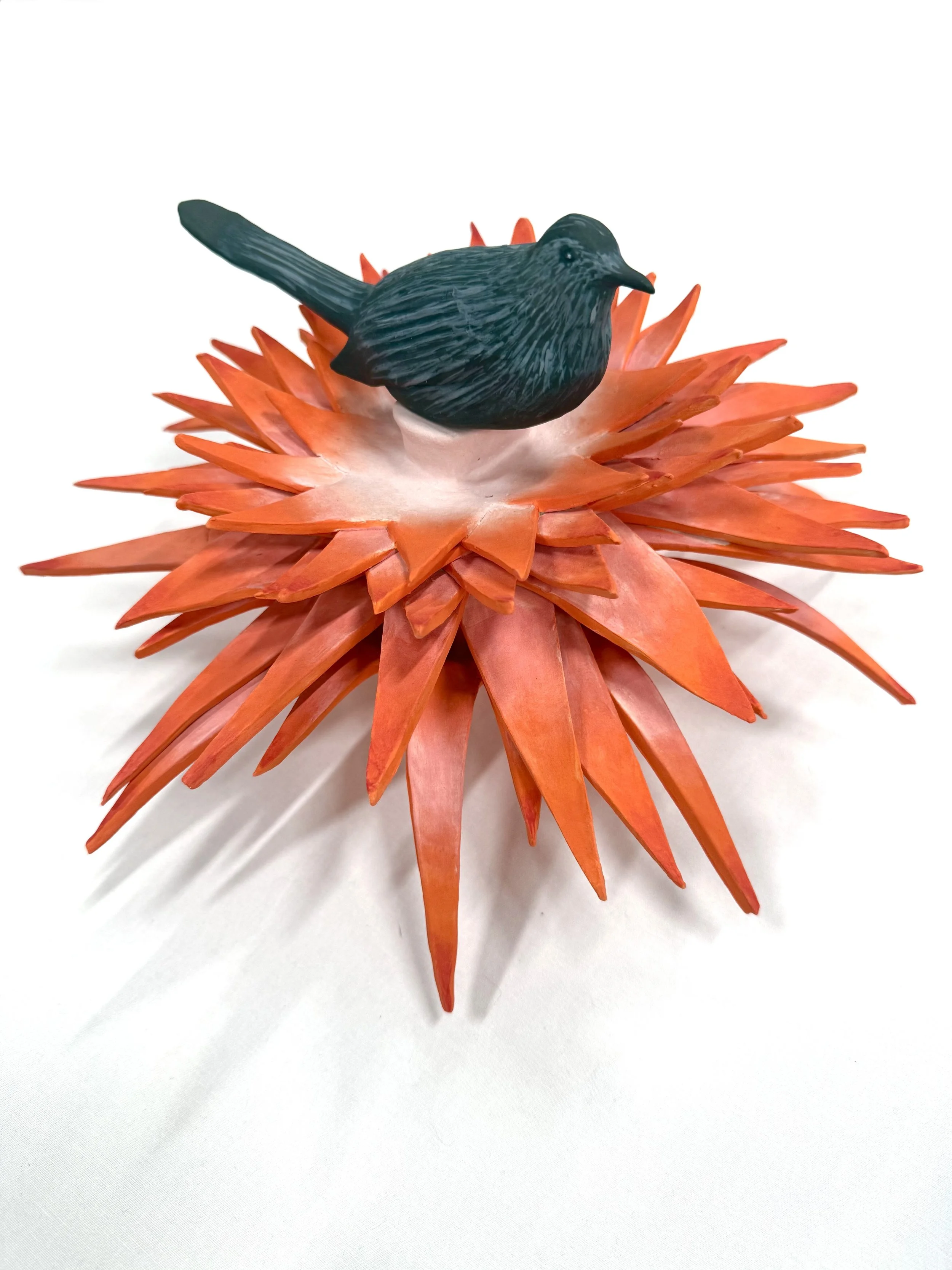 A small black bird figurine perched on a large orange and pink flower crafted from stacked, pointed paper petals.