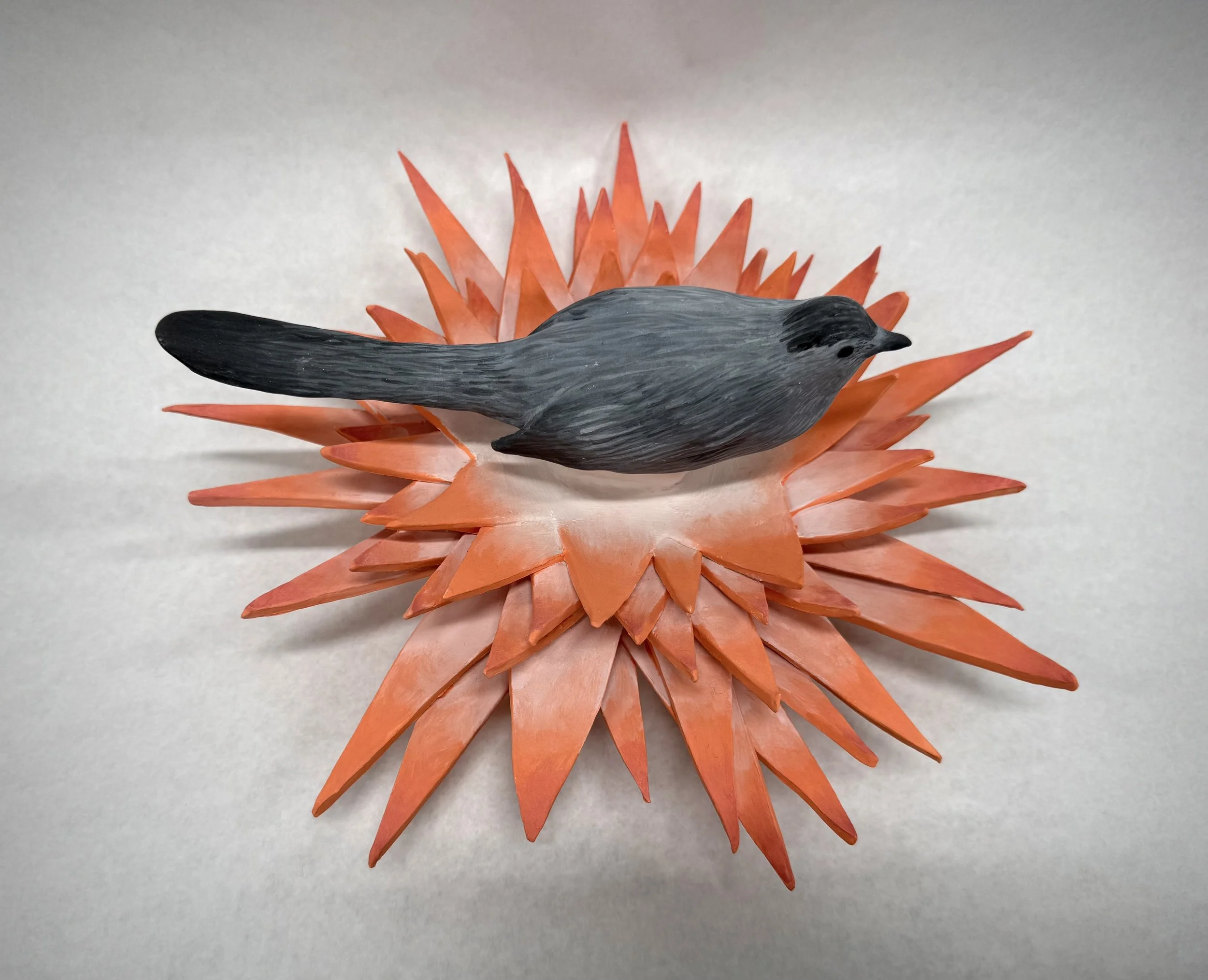 A handcrafted sculpture of a bird with a sleek black body and wings, perched on a vibrant orange spiky base resembling a flower or sunburst.