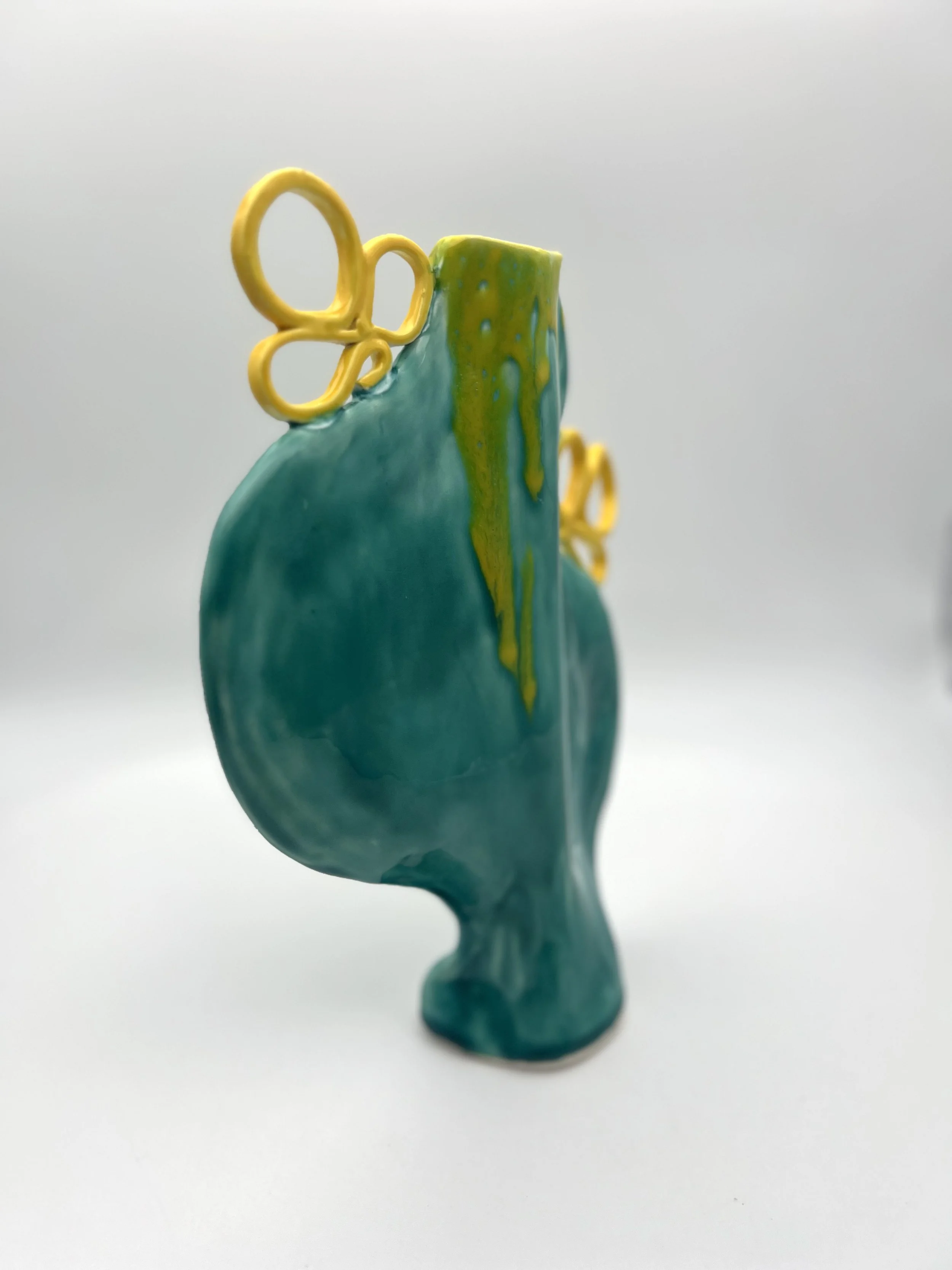 A ceramic sculpture of a humanoid figure with no distinct facial features, colored in shades of blue and green, with yellow looped shapes attached to the top and side, placed on a plain white background.