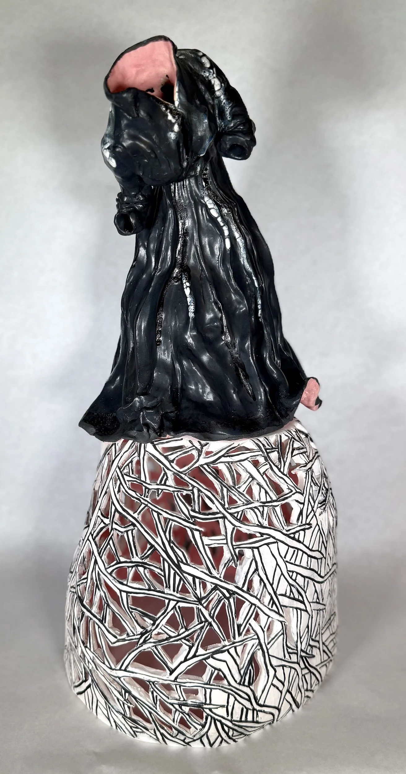 Ceramic sculpture of a shrouded figure with a hollow face, placed on a decorative, web-like patterned base.