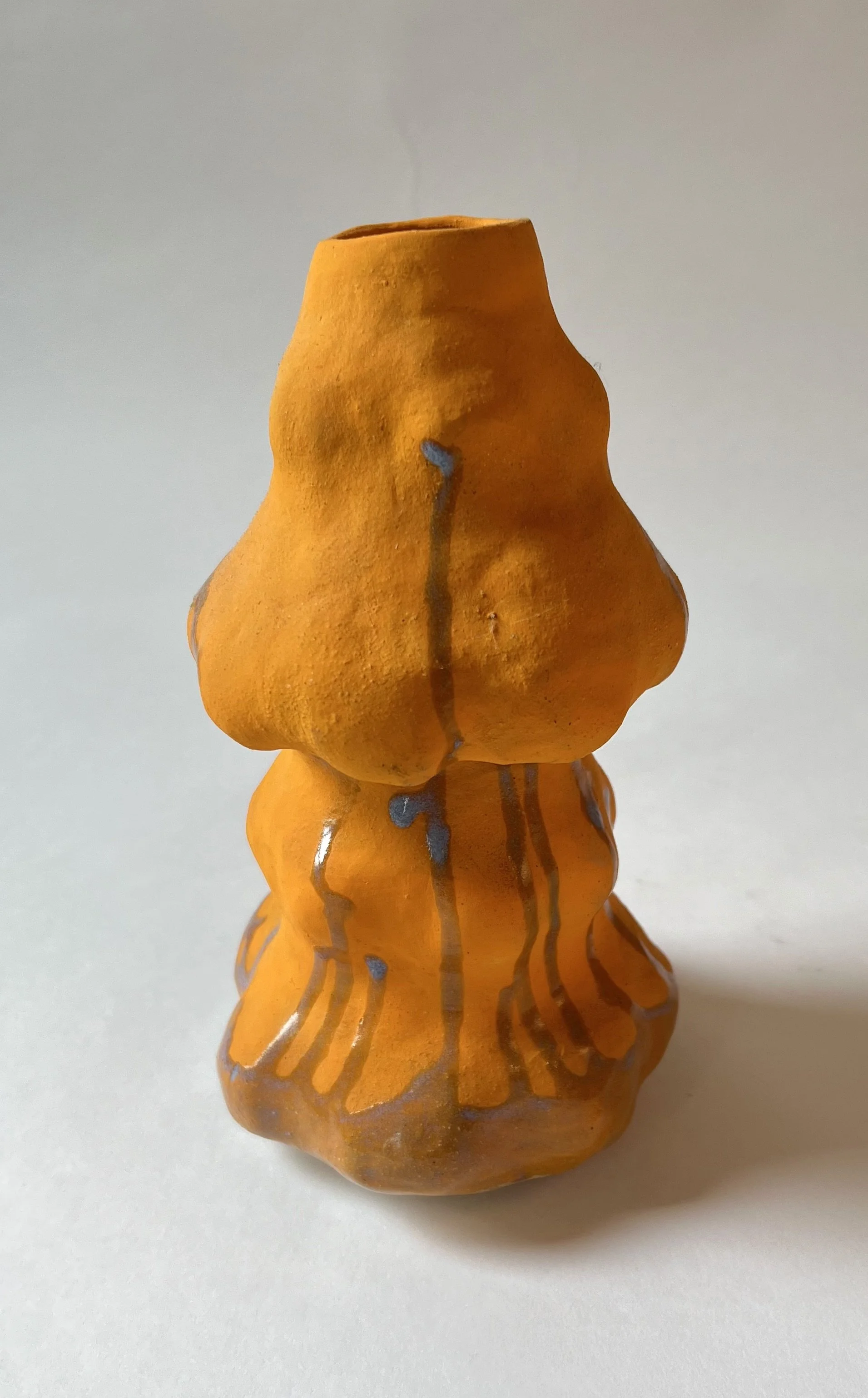 A small, orange ceramic sculpture resembling a life-sized person, with a textured surface and painted silver lines running vertically, set against a plain white background.