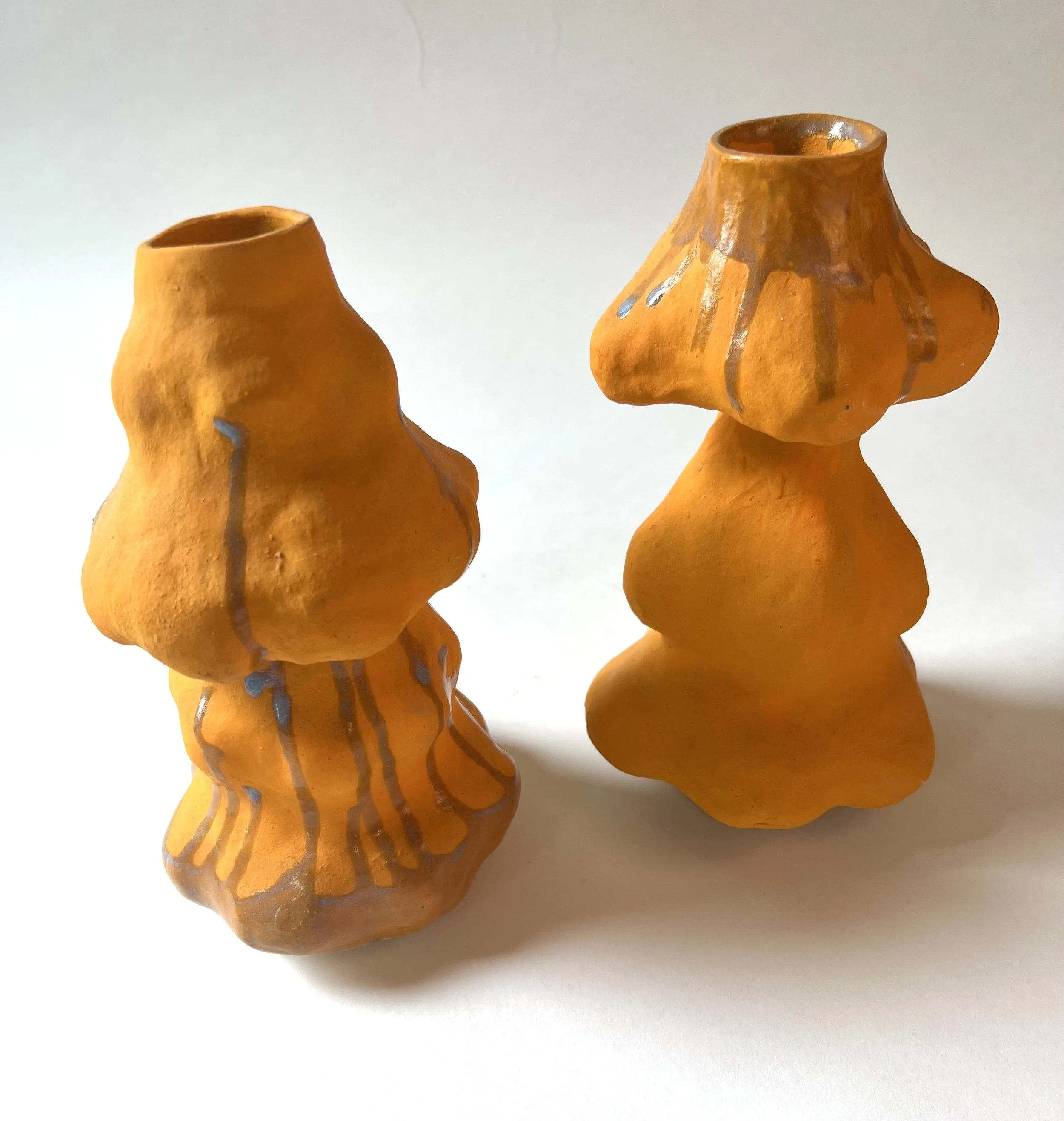 Two orange ceramic vases with irregular shapes and vertical blue and brown streaks are placed on a white surface against a plain white background.