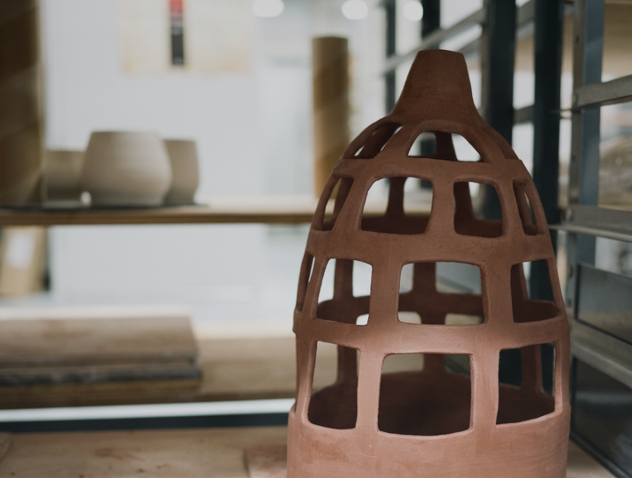 A terracotta decorative sculpture with a grid-like pattern on a wooden surface, with blurred pottery in the background.