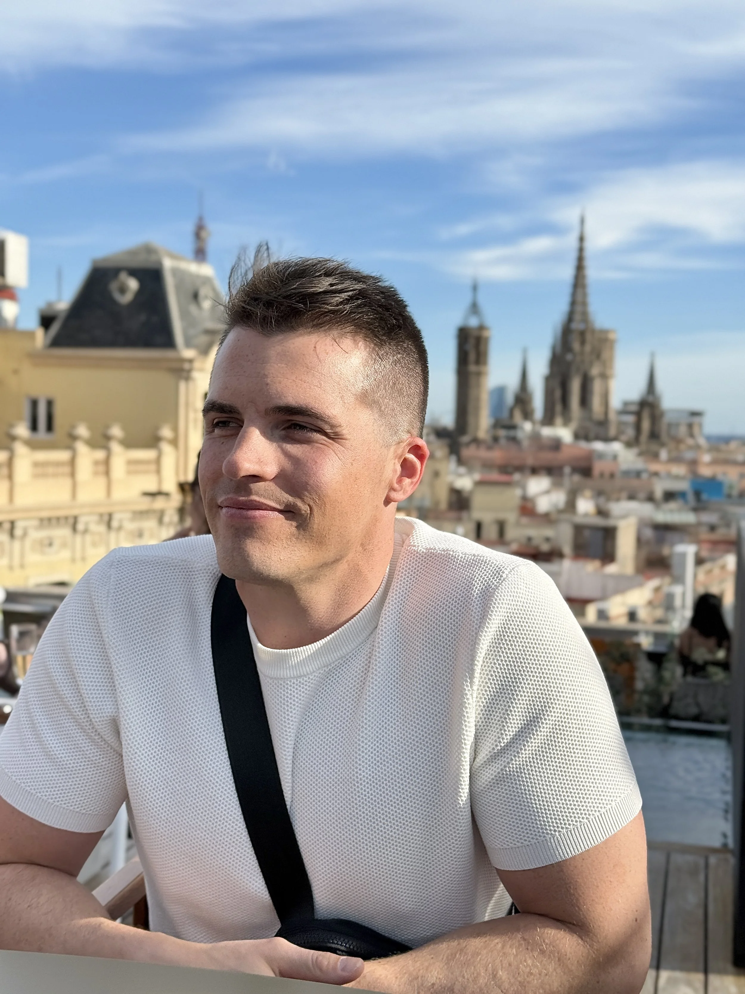 A man sitting outdoors with a city skyline and a church with tall spires in the background.