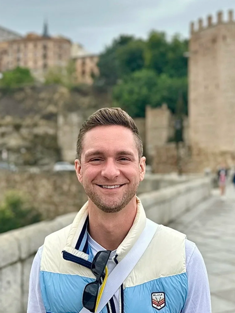 Smiling man standing outdoors on a bridge with historic buildings and a castle in the background.