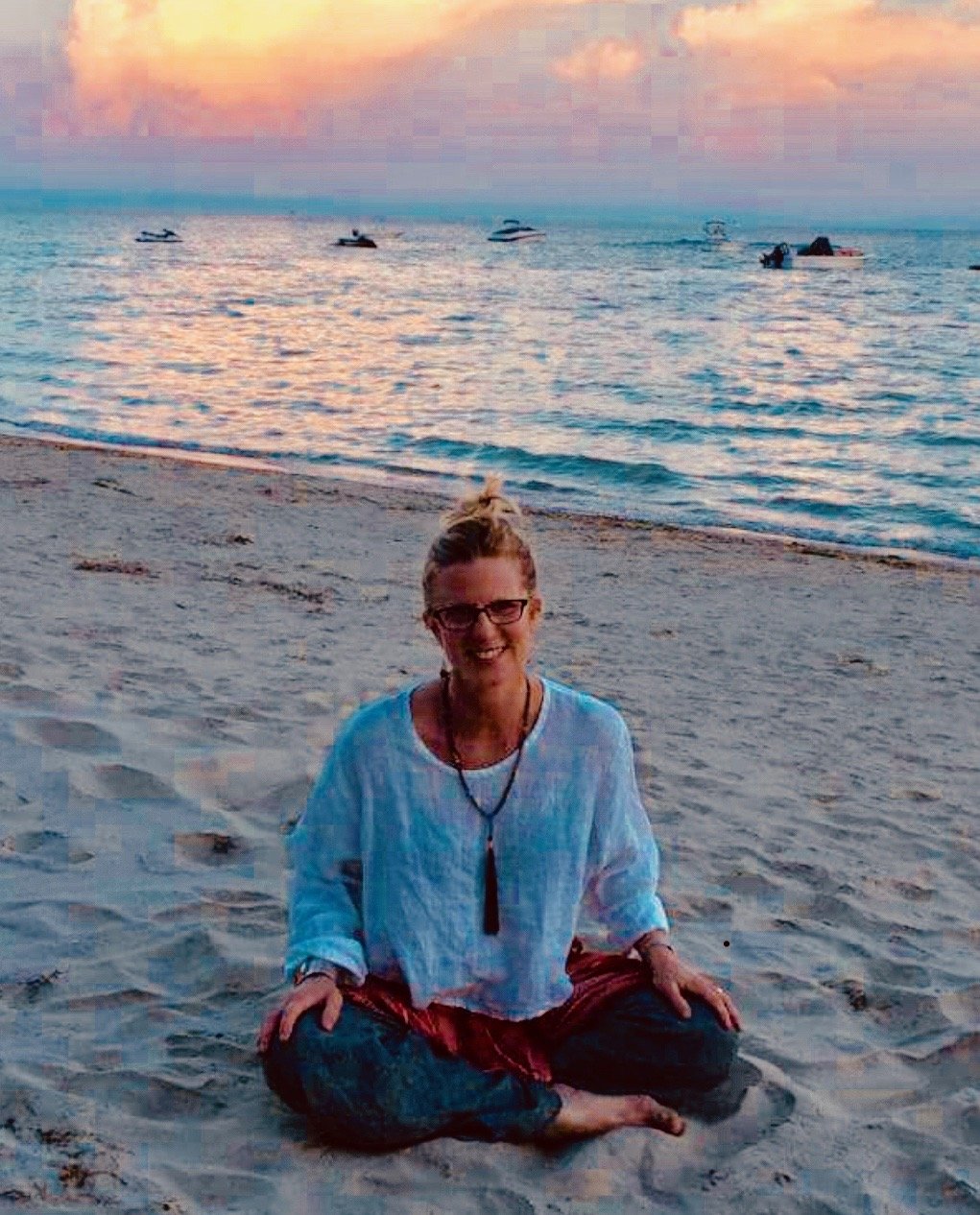 A smiling woman with glasses sitting cross-legged on the sandy beach at sunset, with boats floating on the water in the background.