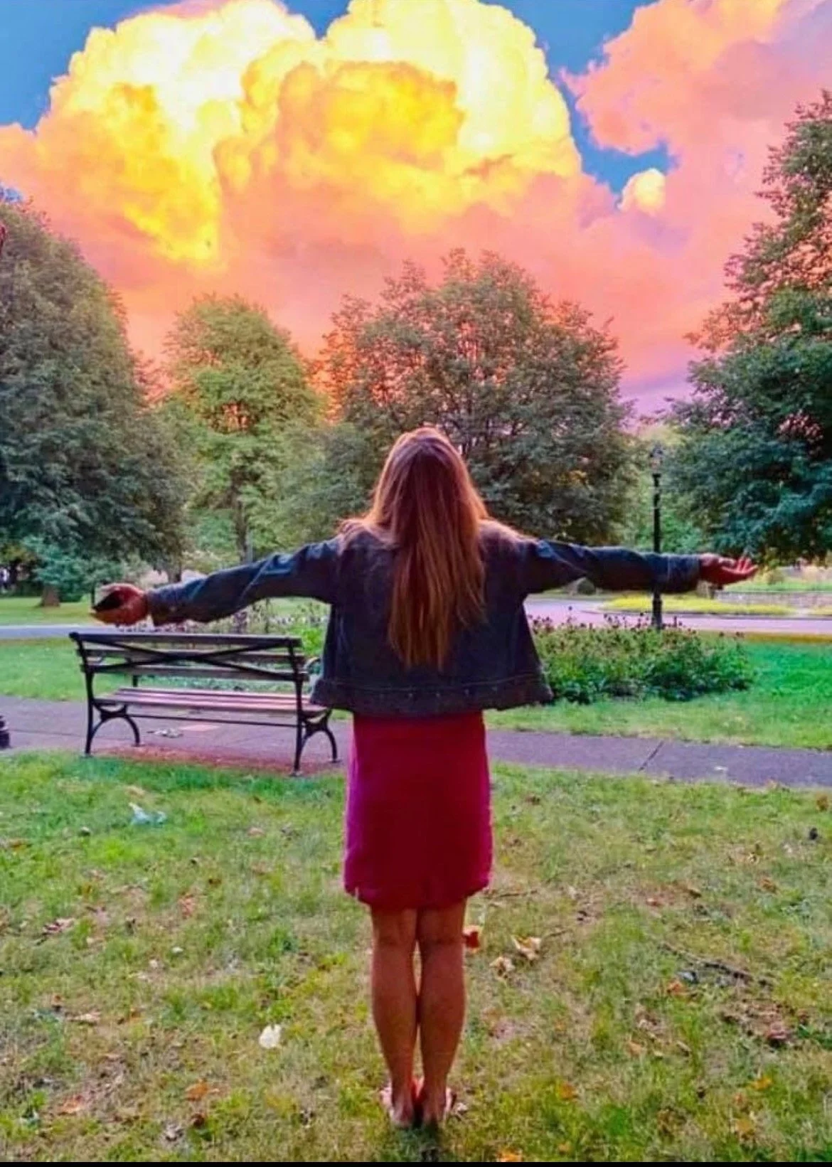 A woman with long hair in a pink skirt and black jacket stands in a park with her arms outstretched. The sky is filled with vibrant pink, orange, and yellow clouds, creating a dramatic sunset or sunrise scene.