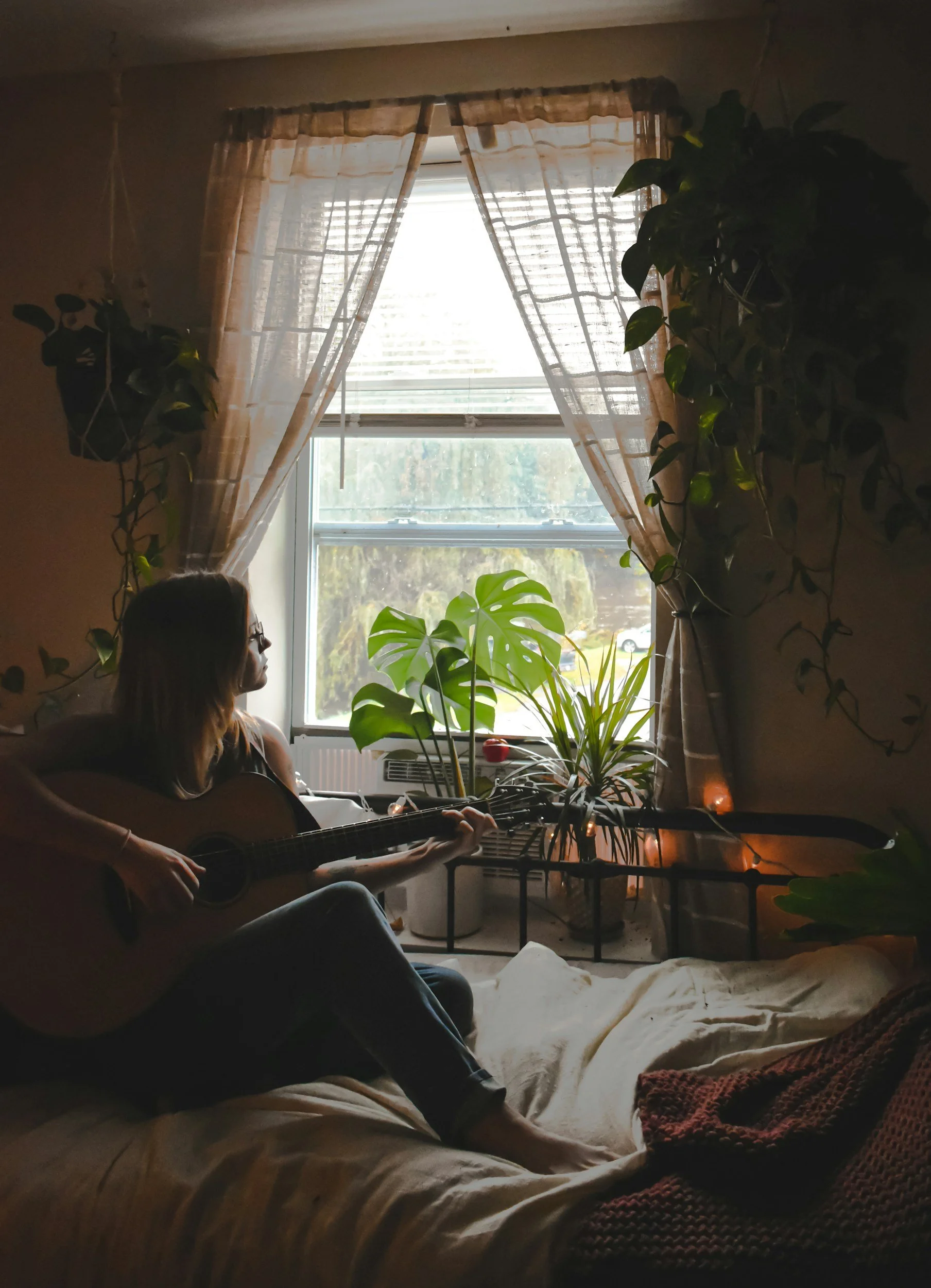 A woman with glasses and long hair sitting on a bed with cream-colored blankets, playing an acoustic guitar. The room has large windows with sheer curtains, several green plants on the windowsill and beside the window, and string lights at the edge of the bed.