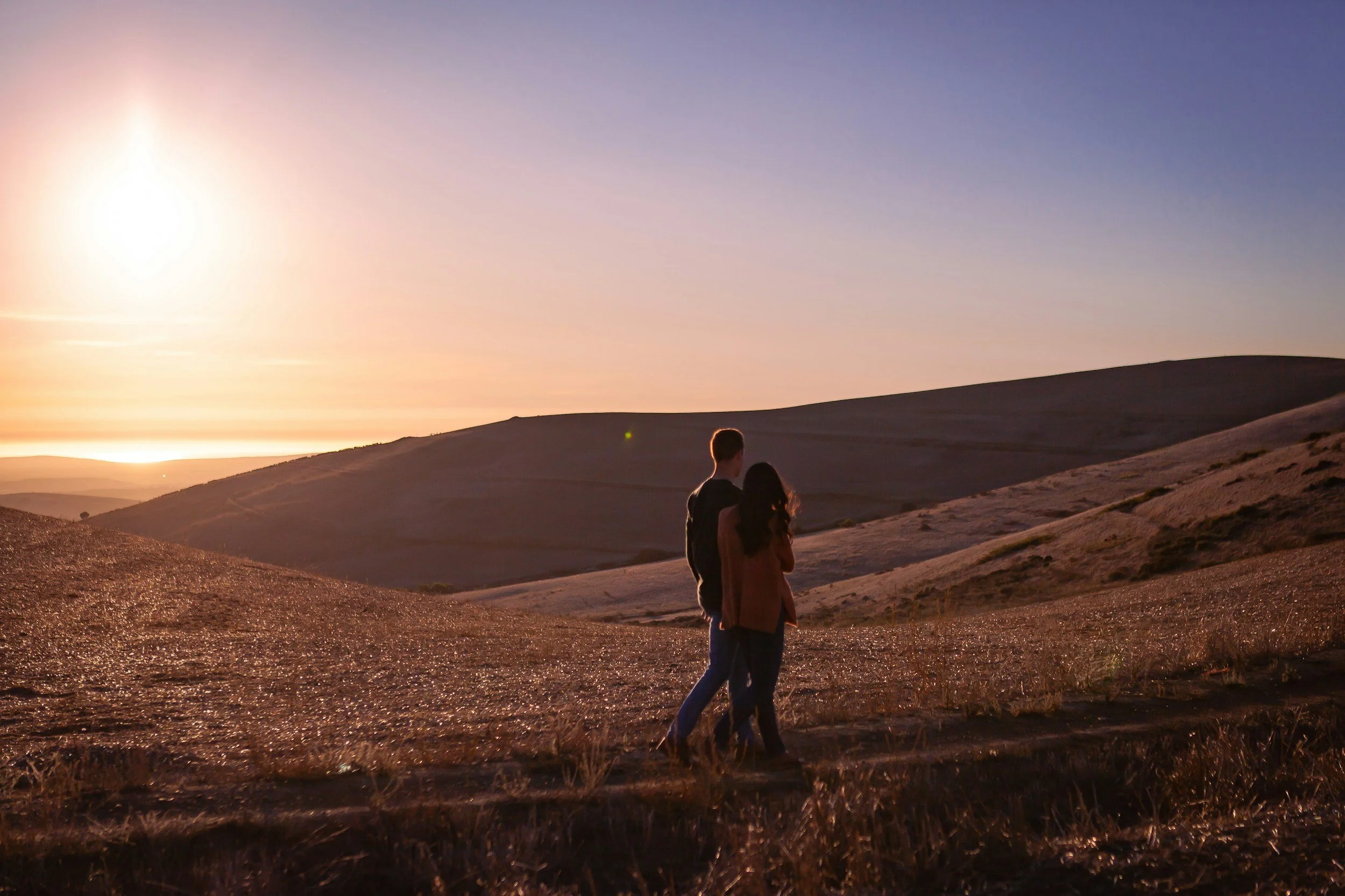 A couple walking in a desert landscape during sunset or sunrise with hills and a clear sky.