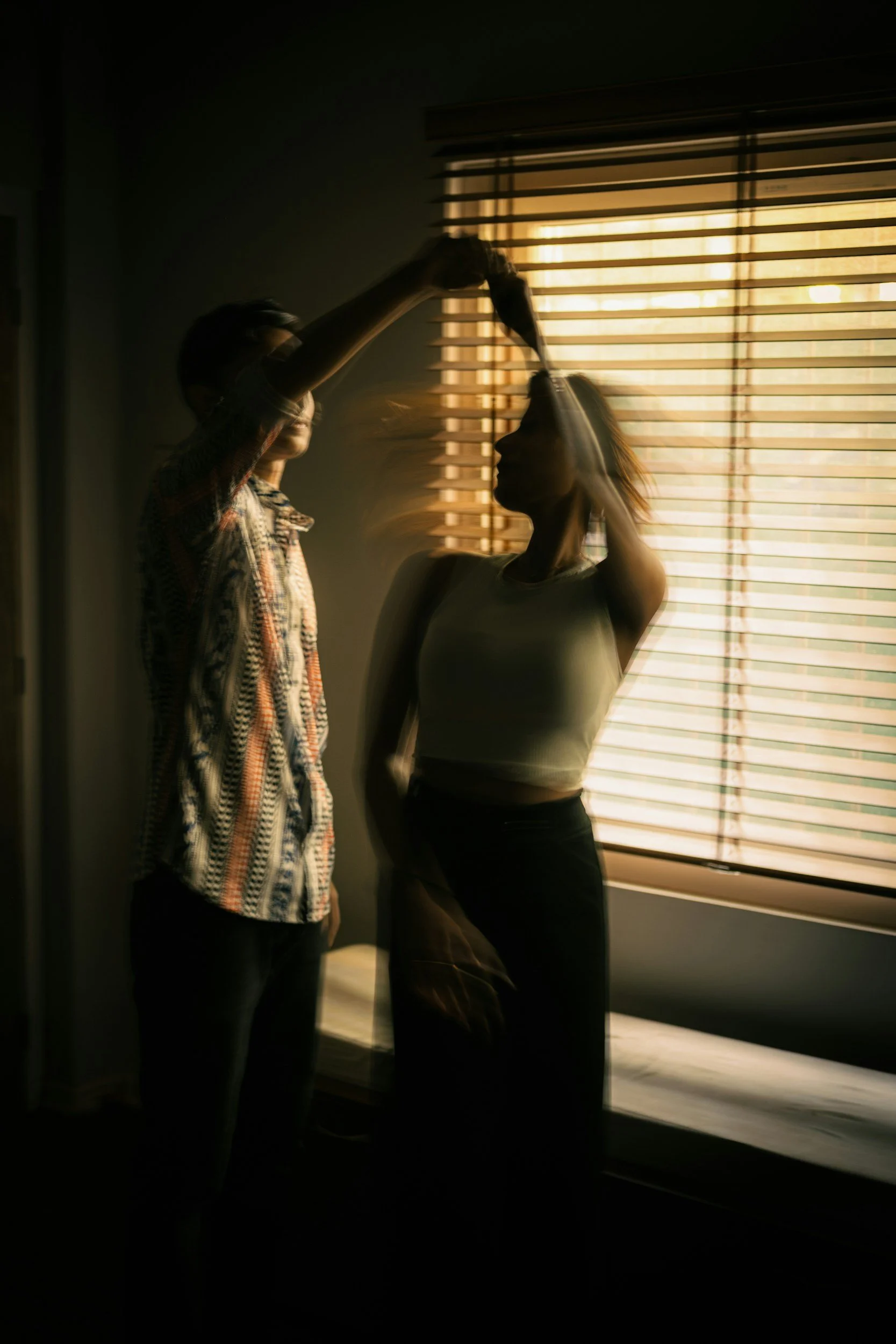 Two women standing in front of a window with horizontal blinds, one pouring a drink into the other's mouth during sunset.