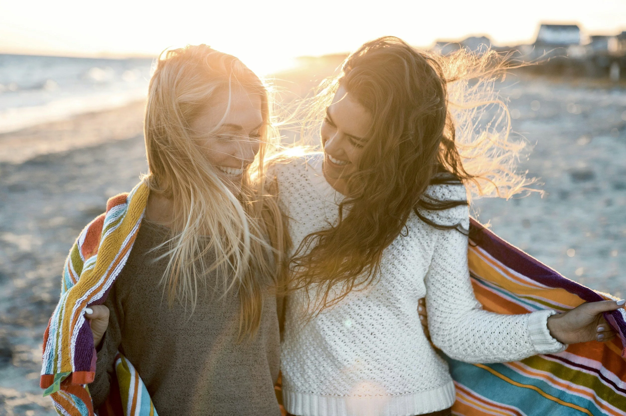 Two smiling women with long hair sharing a moment at the beach during sunset, one wrapped in a colorful towel.