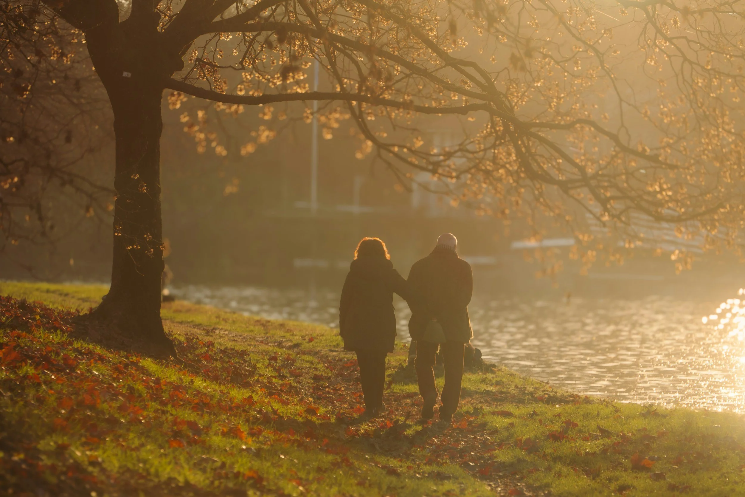 Two people walking along a lakeside path with trees and autumn foliage, during sunset or sunrise, creating a warm and peaceful atmosphere.