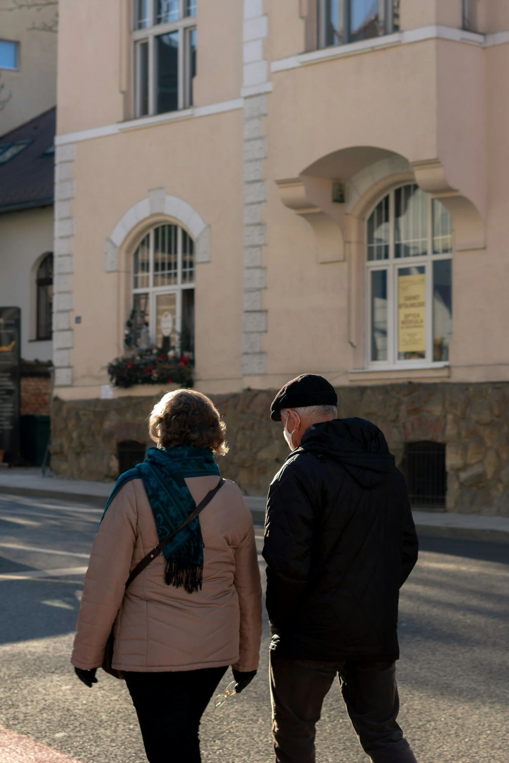 Two people walking down a city street, viewed from behind. One wears a beige coat and green scarf, the other is in a black jacket and black beret, with their heads close together. In the background, a beige building with arched windows.