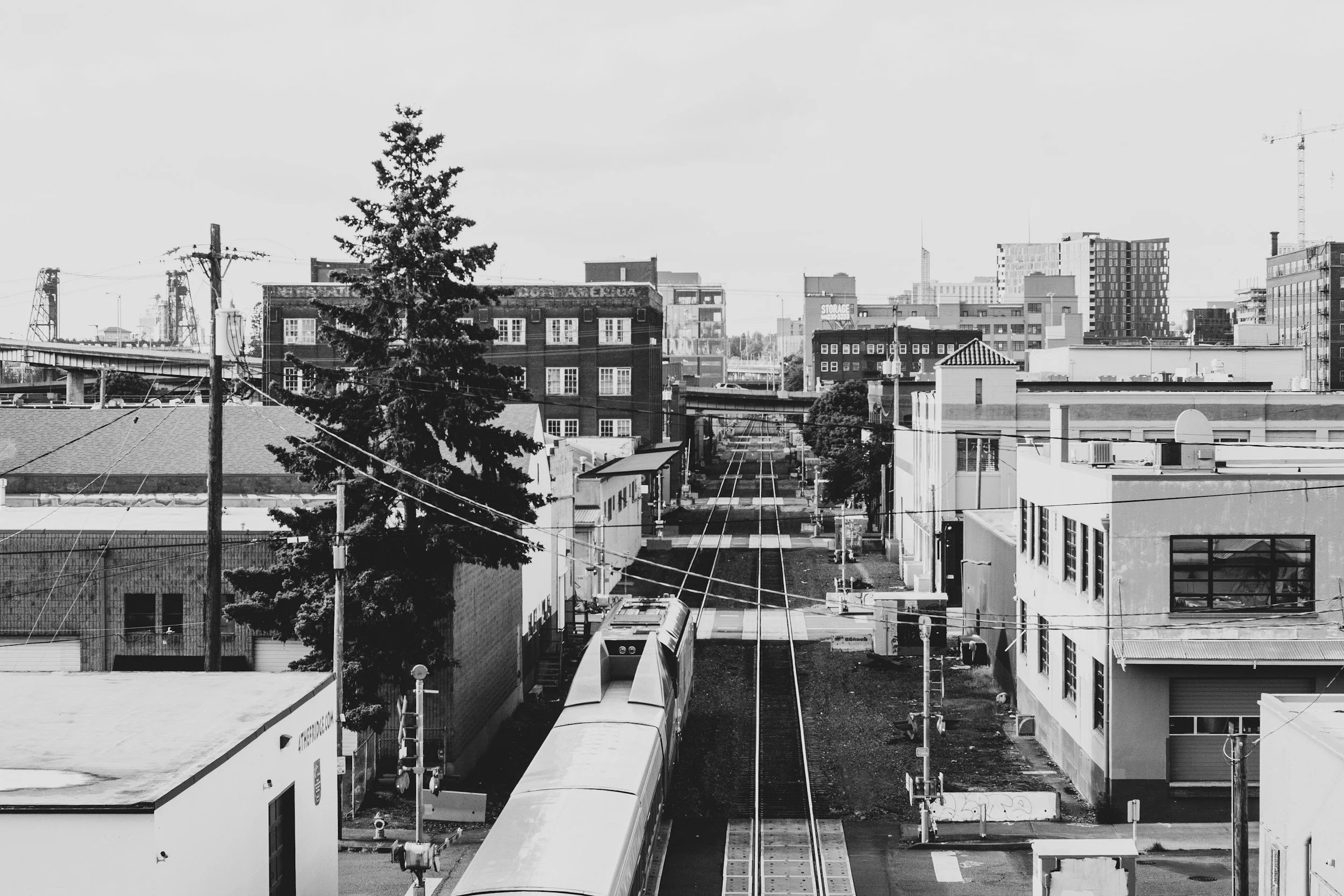 A black-and-white photo of an urban train station with train tracks, train cars, and surrounding buildings and power lines.