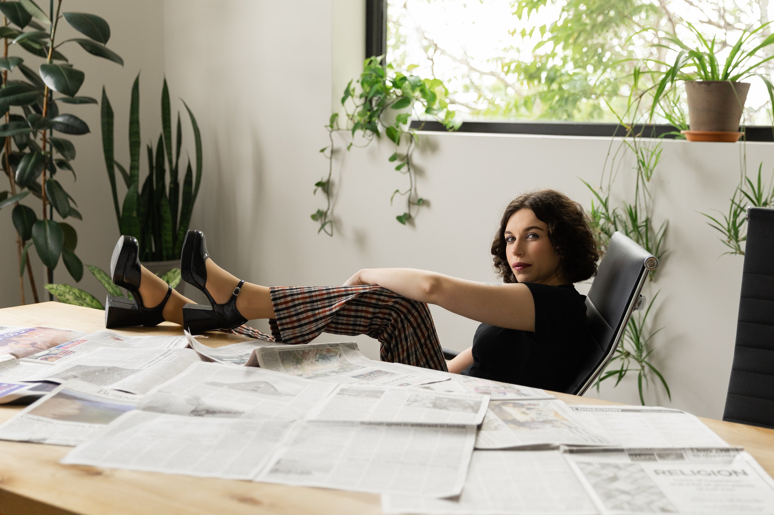 A woman with dark curly hair and wearing a black shirt, plaid pants, and black high heels, is sitting in a black office chair with her legs extended on a table covered with newspapers. The room has white walls and large windows with several potted green plants. The woman is looking at the camera with a relaxed expression.