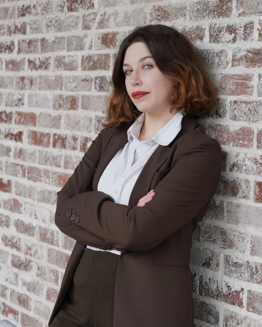 A woman with wavy brown hair and light skin, dressed in a white collared shirt and dark brown blazer, standing with crossed arms against a brick wall, looking confidently at the camera.