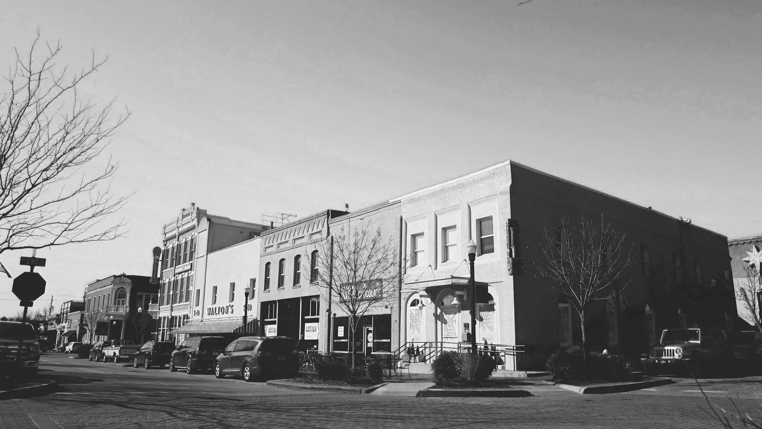 Black and white photo of a small downtown shopping district with parked cars, leafless trees, and buildings including Valvol's and Chocolate stores.
