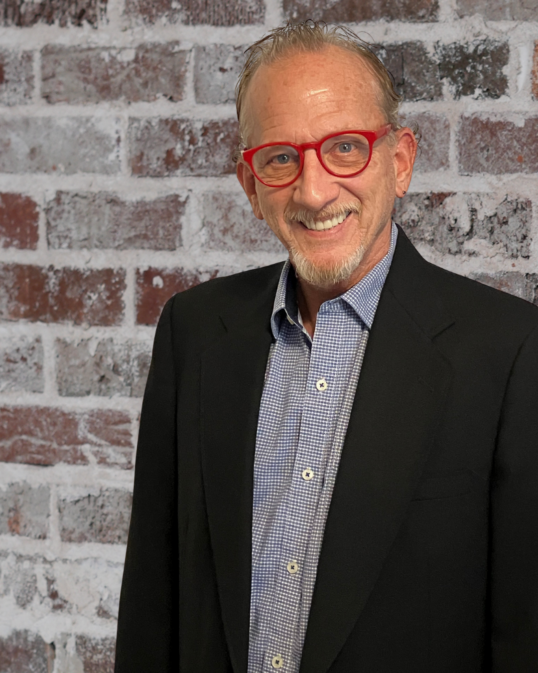 A smiling middle-aged man with red glasses, wearing a black blazer and a blue checkered shirt, standing in front of a brick wall.