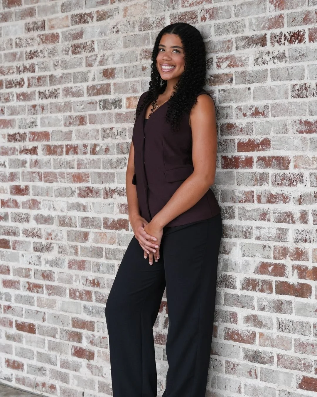 A woman standing and smiling against a brick wall, wearing a dark sleeveless top and black pants.