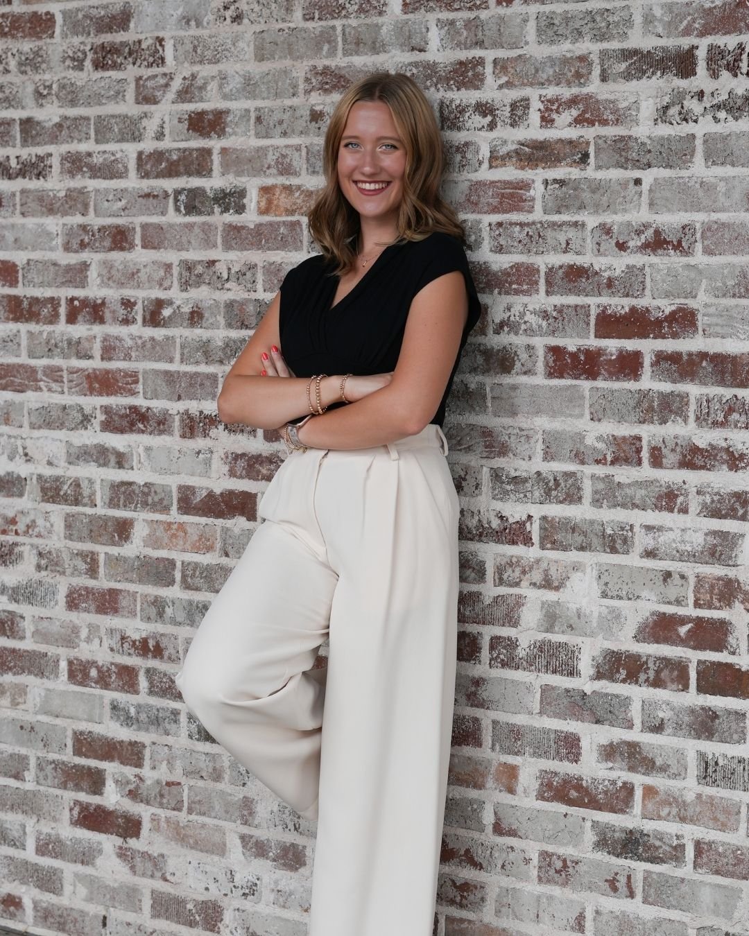 A woman stands with arms crossed against a brick wall, wearing a black top and white high-waisted pants, smiling at the camera.