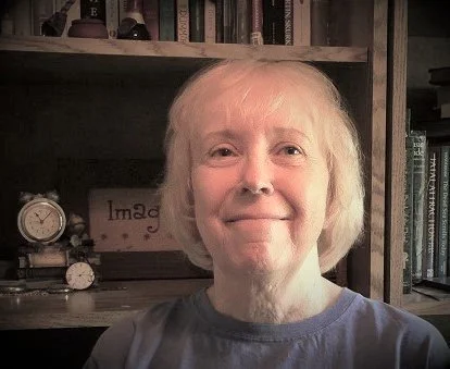 A close-up of an woman with short, light-colored hair smiling at the camera, with a bookshelf and clock in the background.