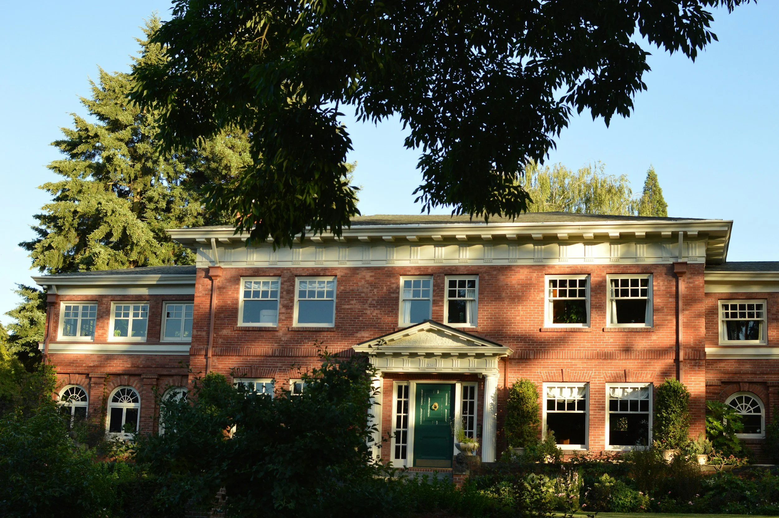 A large brick house with a green door surrounded by trees and plants under a clear blue sky.
