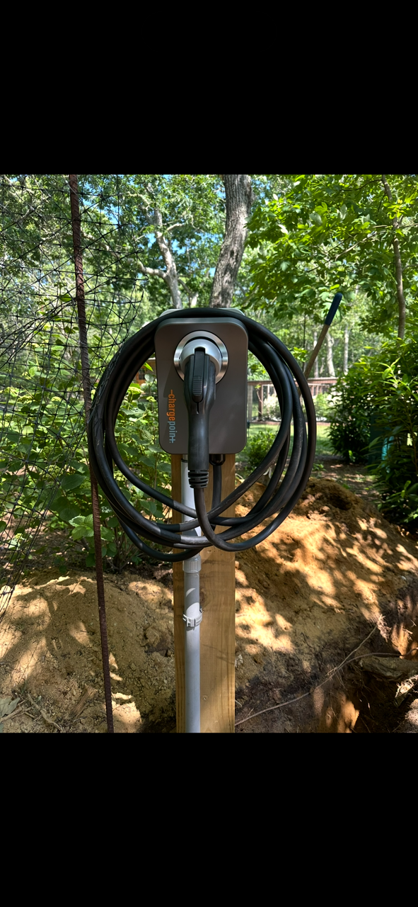 Electric vehicle charging station in a wooded outdoor setting with trees and plants, mounted on a wooden post.