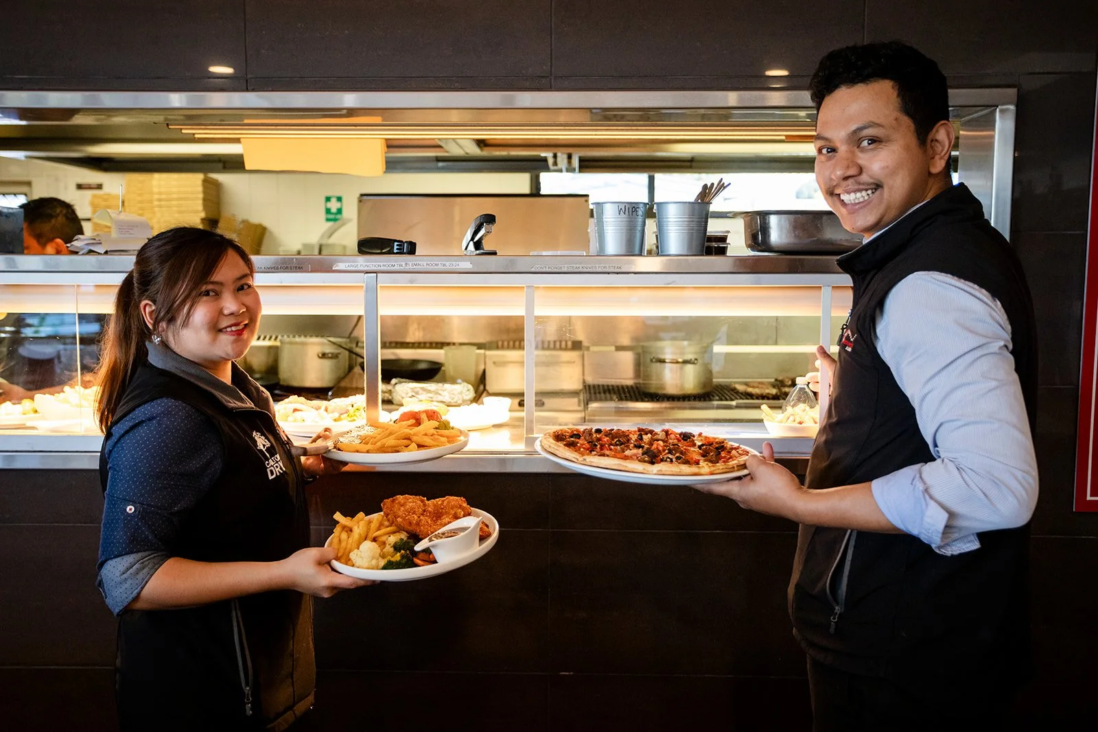Two restaurant workers, a woman and a man, smiling and holding plates of food in a restaurant kitchen. The woman holds a plate with fries, cauliflower, and chicken, while the man shows a pizza.