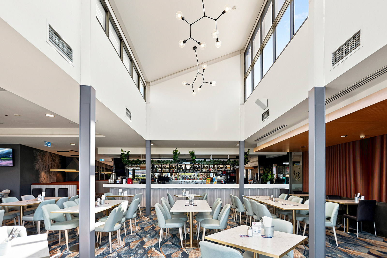 Interior of a modern restaurant or cafe with light-colored chairs, white tables, and a stylish chandelier hanging from the high ceiling. Large windows provide natural light, and a bar area with plants is visible in the background.