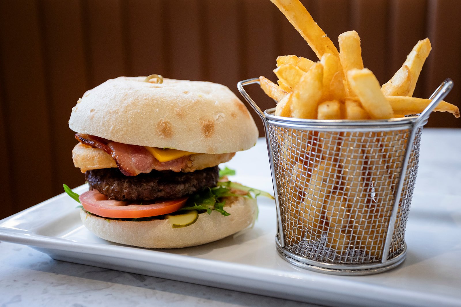 A cheeseburger with bacon and vegetables next to a basket of French fries on a white plate.
