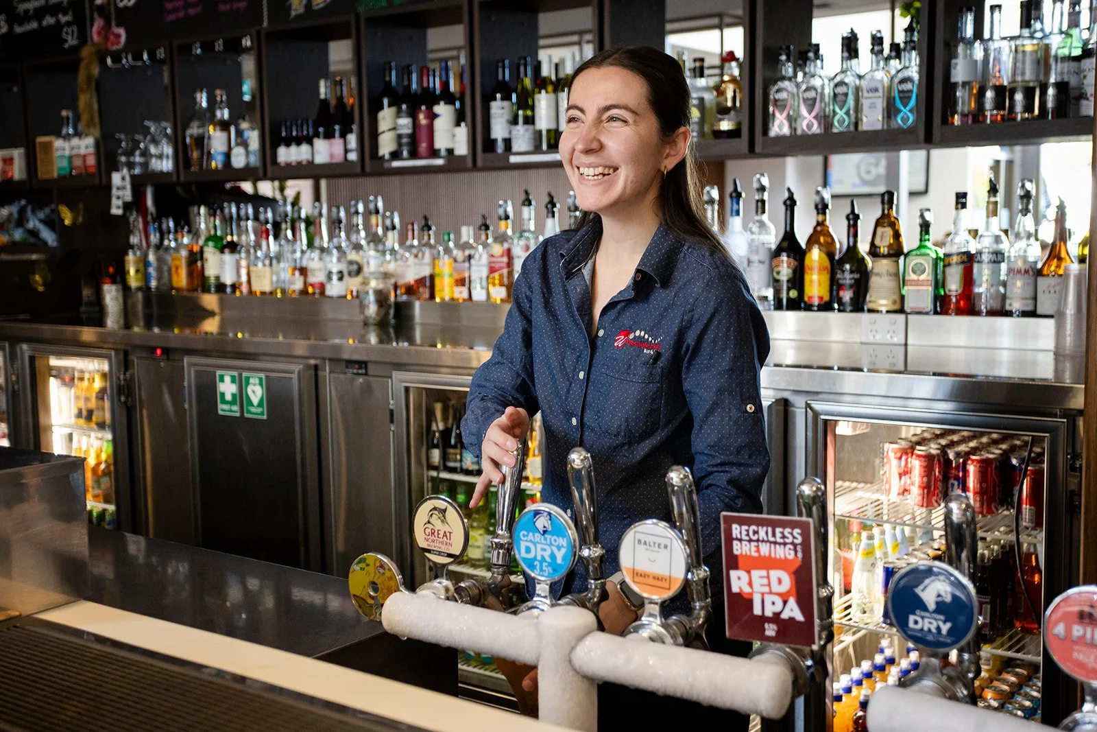 A woman with dark hair smiling behind a bar counter with beverage taps, bottles of alcohol in the background, and various signs for beer brands.