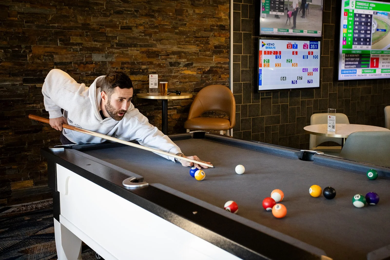 A man playing pool at a billiards table in a bar or pub, leaning over and aiming to hit the balls with a cue stick, with digital screens showing betting odds and horse race results in the background.