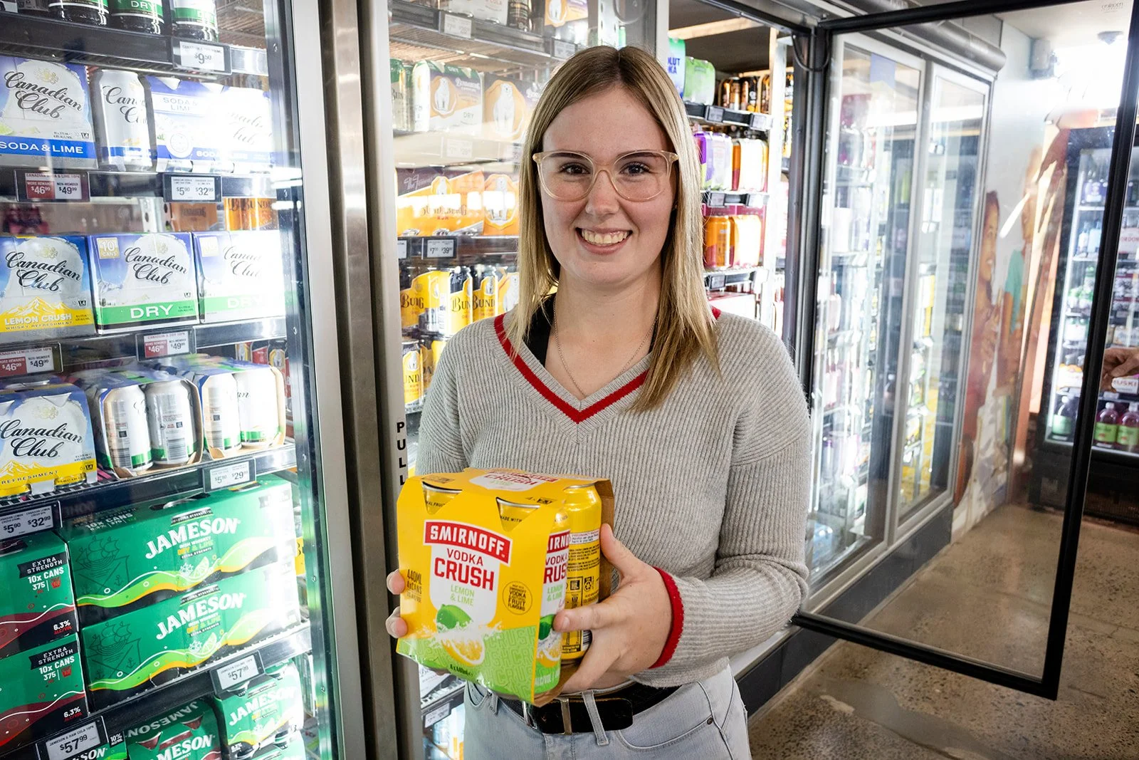 A woman with glasses and a striped sweater holding a six-pack of Smirnoff Vodka Crush Lemon Lime in front of a convenience store refrigerated section.