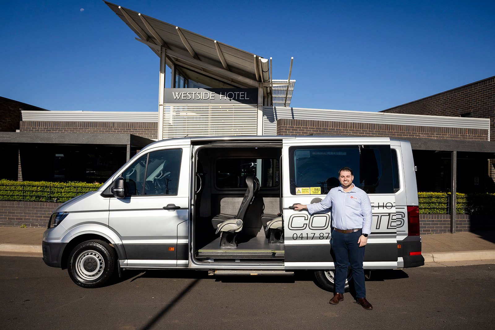 A smiling man standing outside a silver shuttle bus in front of the Westside Hotel on a sunny day.