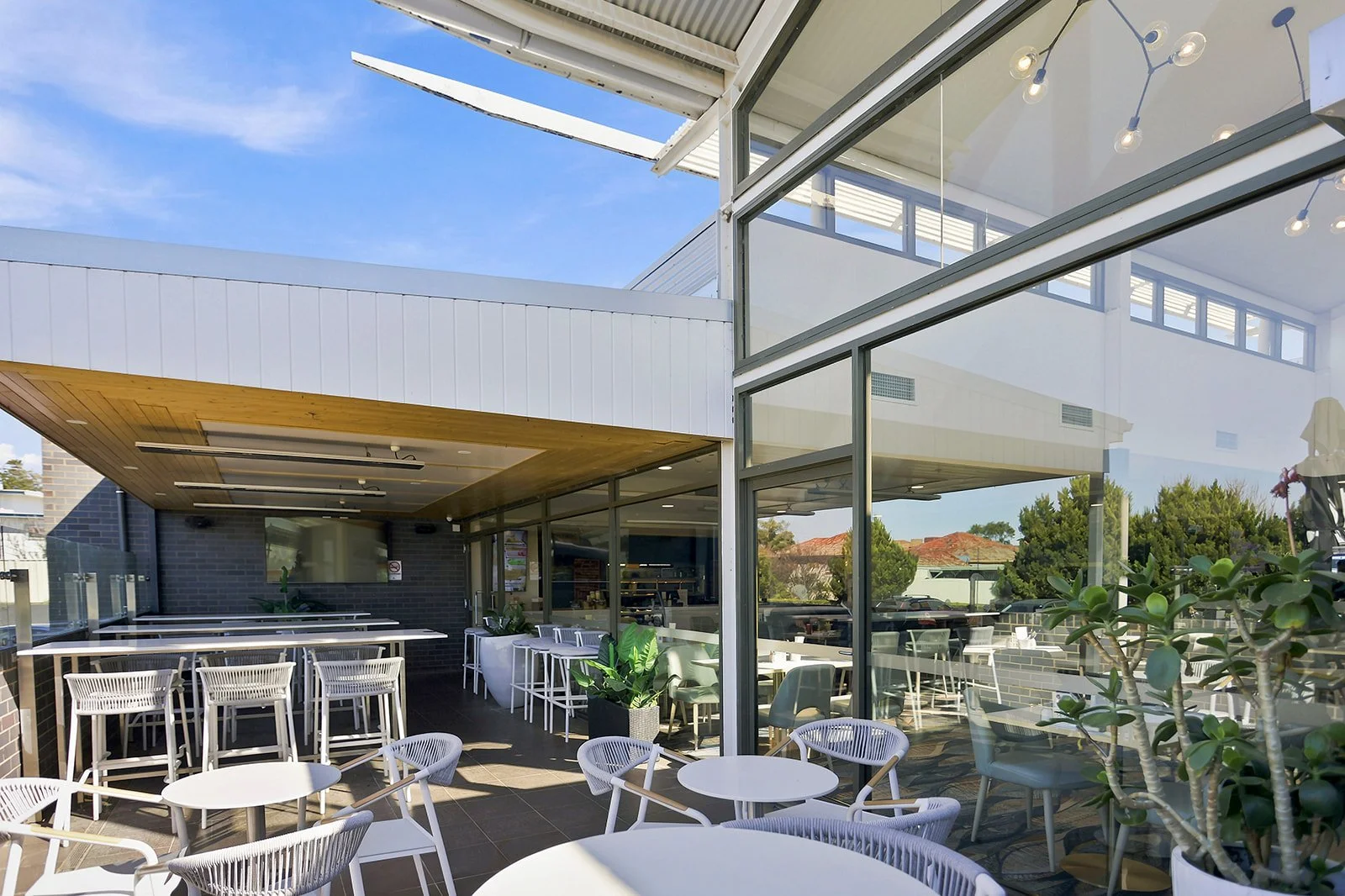 Outdoor patio seating area with white chairs and tables, surrounded by glass walls, greenery, and part of a modern building with glass and wood details, under a blue sky.