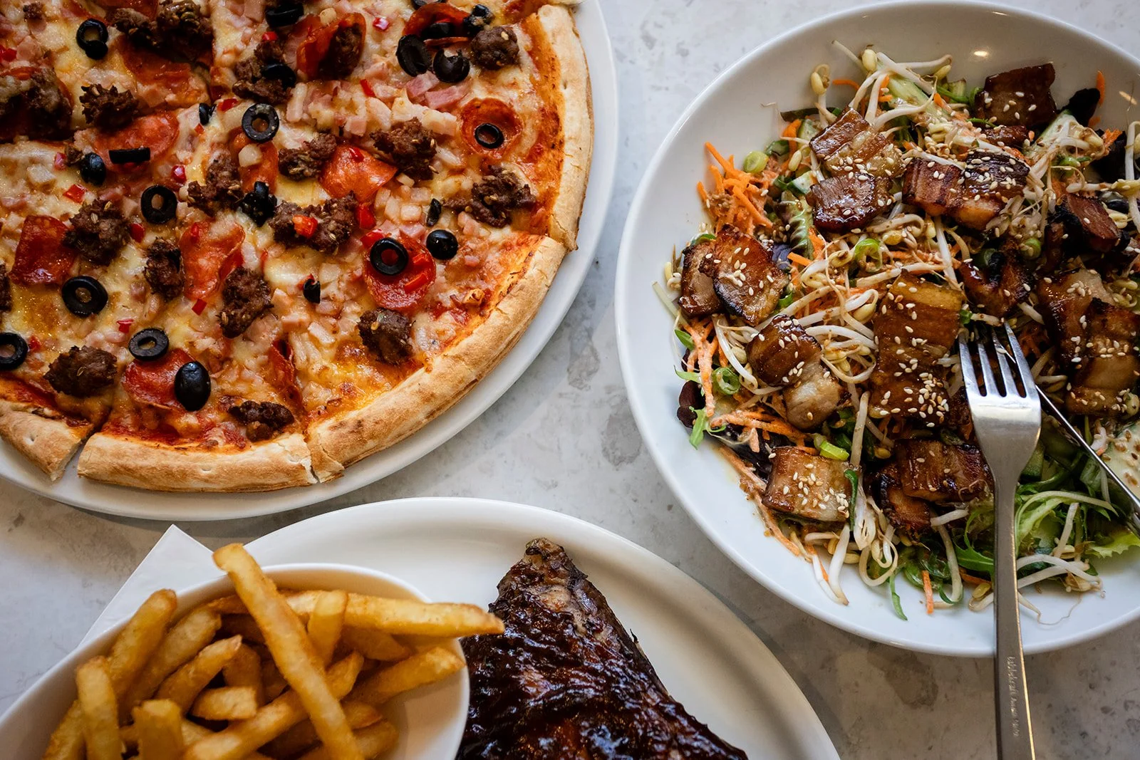 A meal consisting of a pizza with pepperoni, sausage, black olives, and cheese, a salad with tofu and vegetables topped with sesame seeds, a bowl of French fries, and a rack of ribs with barbecue sauce on a marble table.
