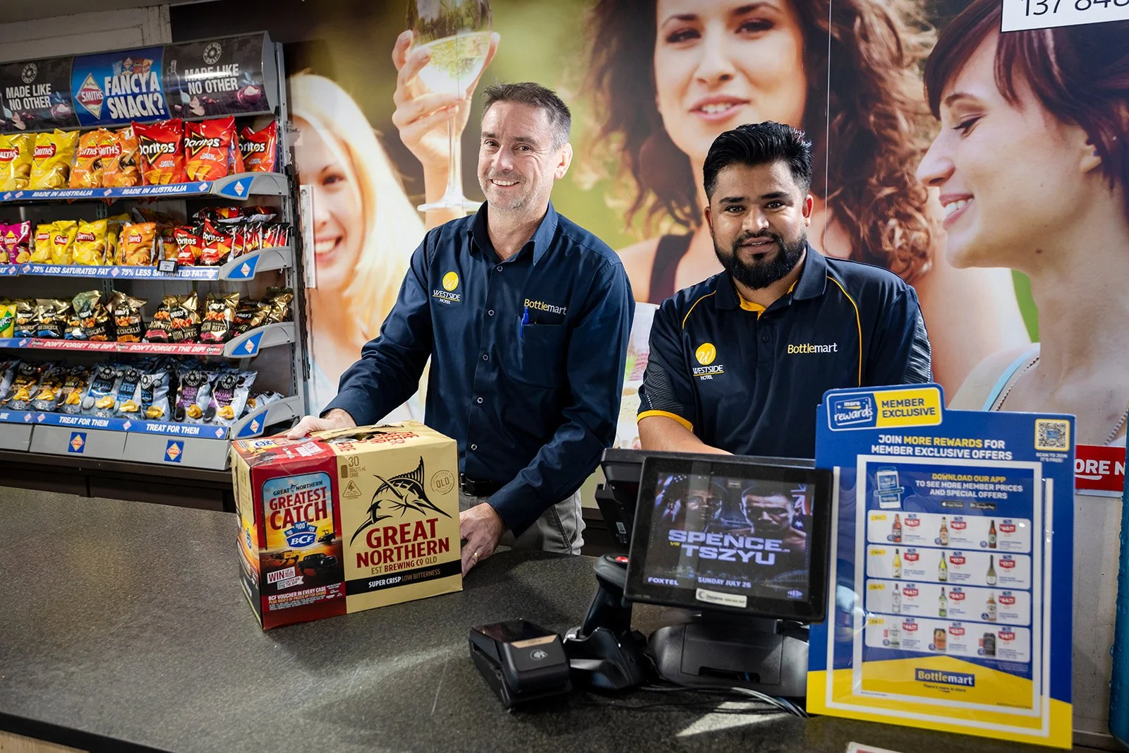 Two men working at a checkout counter in a store, with snack shelves behind them and promotional displays on the counter.