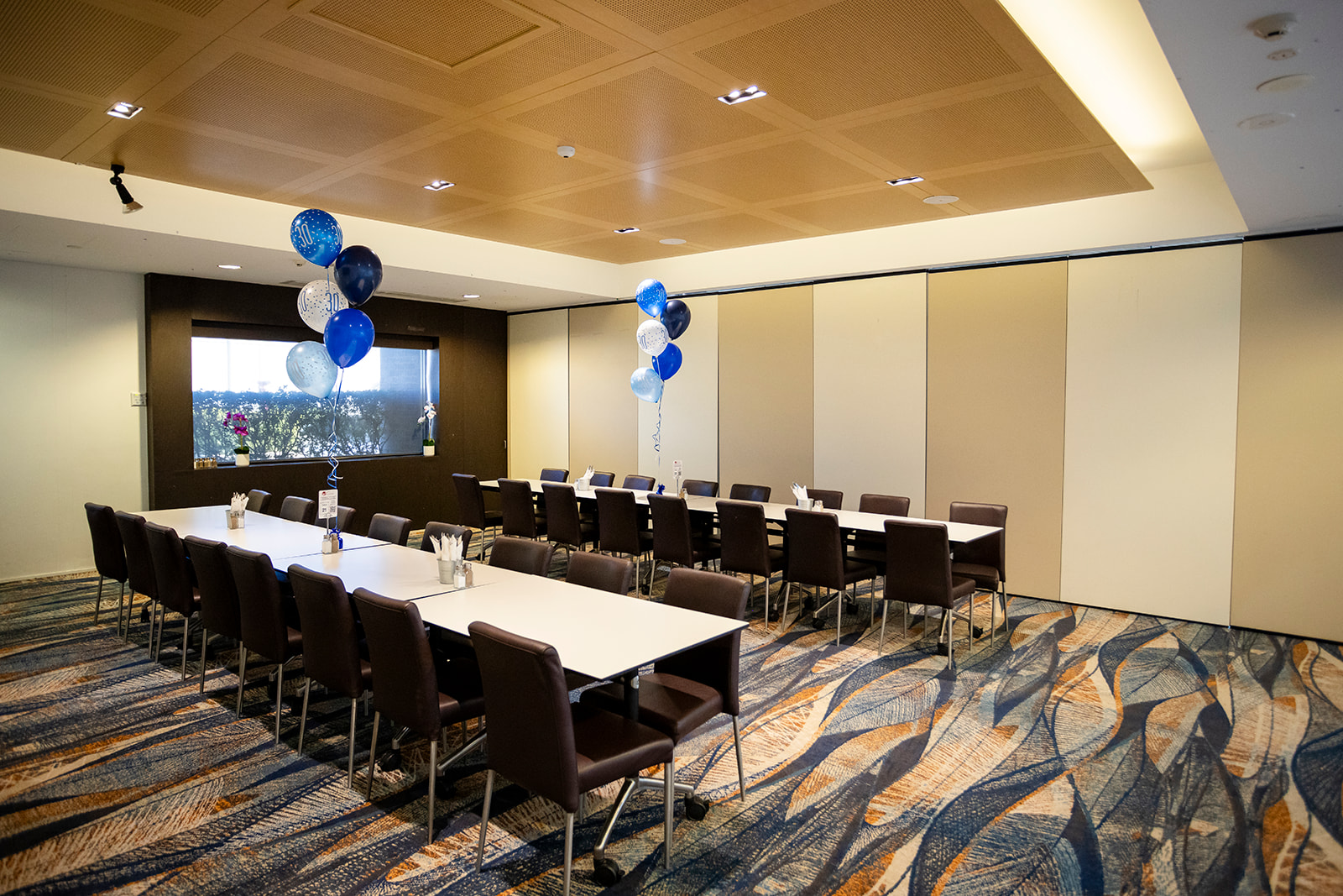 A decorated conference room with long white tables, black chairs, blue and white balloons, and a large window with potted orchids inside.