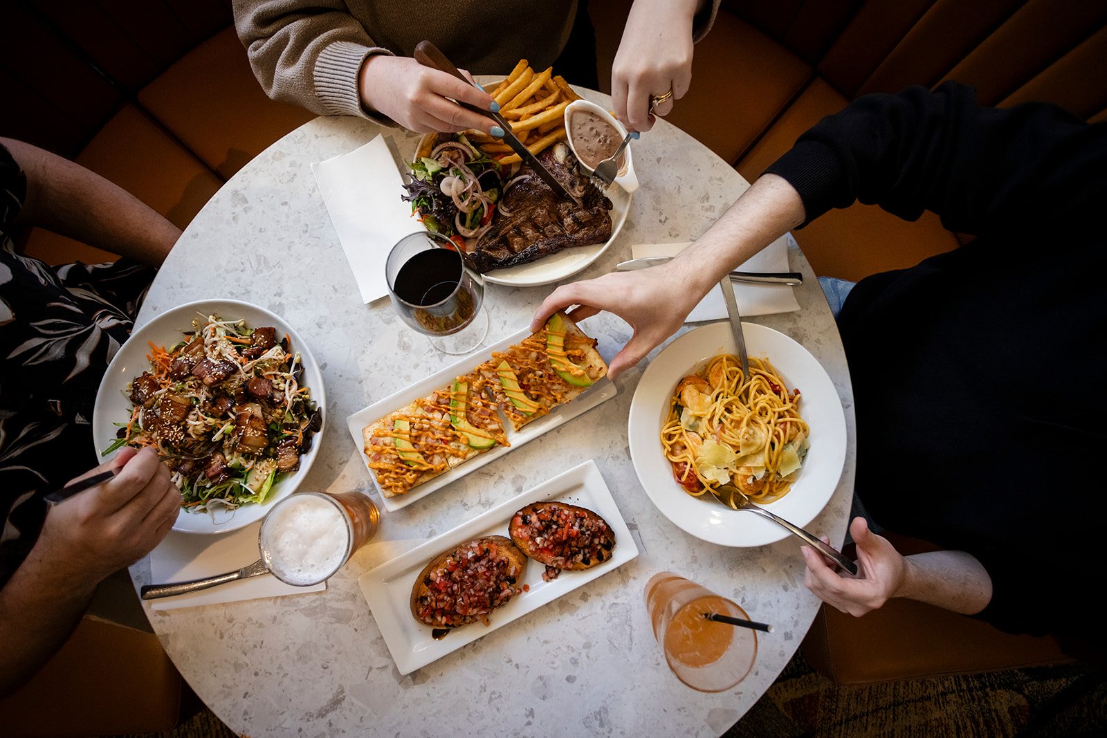 Top-down view of a round table with four people sharing a meal, including a large steak with sides, pasta, a salad, and drinks.