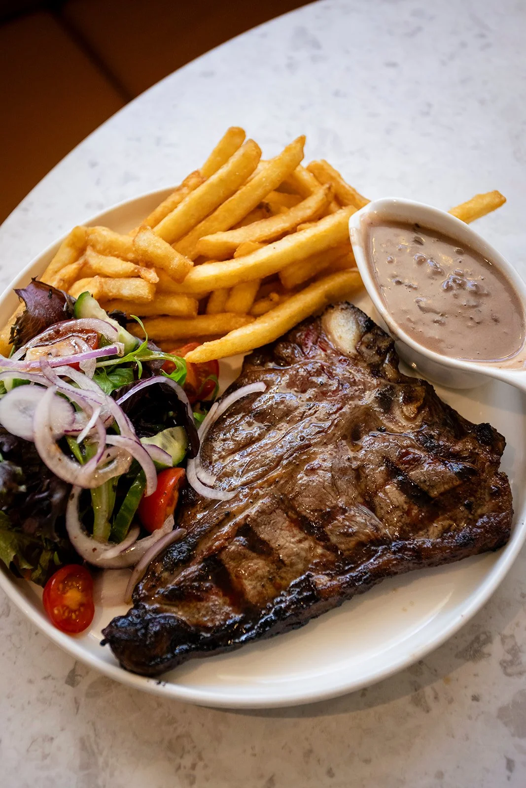 Plate with grilled steak, French fries, fresh salad with cherry tomatoes, and a bowl of gravy.